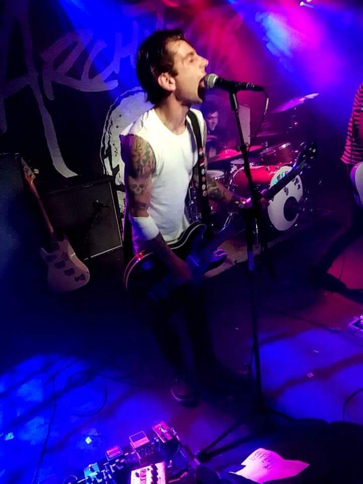 Musician singing into a microphone on stage with colorful lighting, wearing a sleeveless shirt and playing guitar.