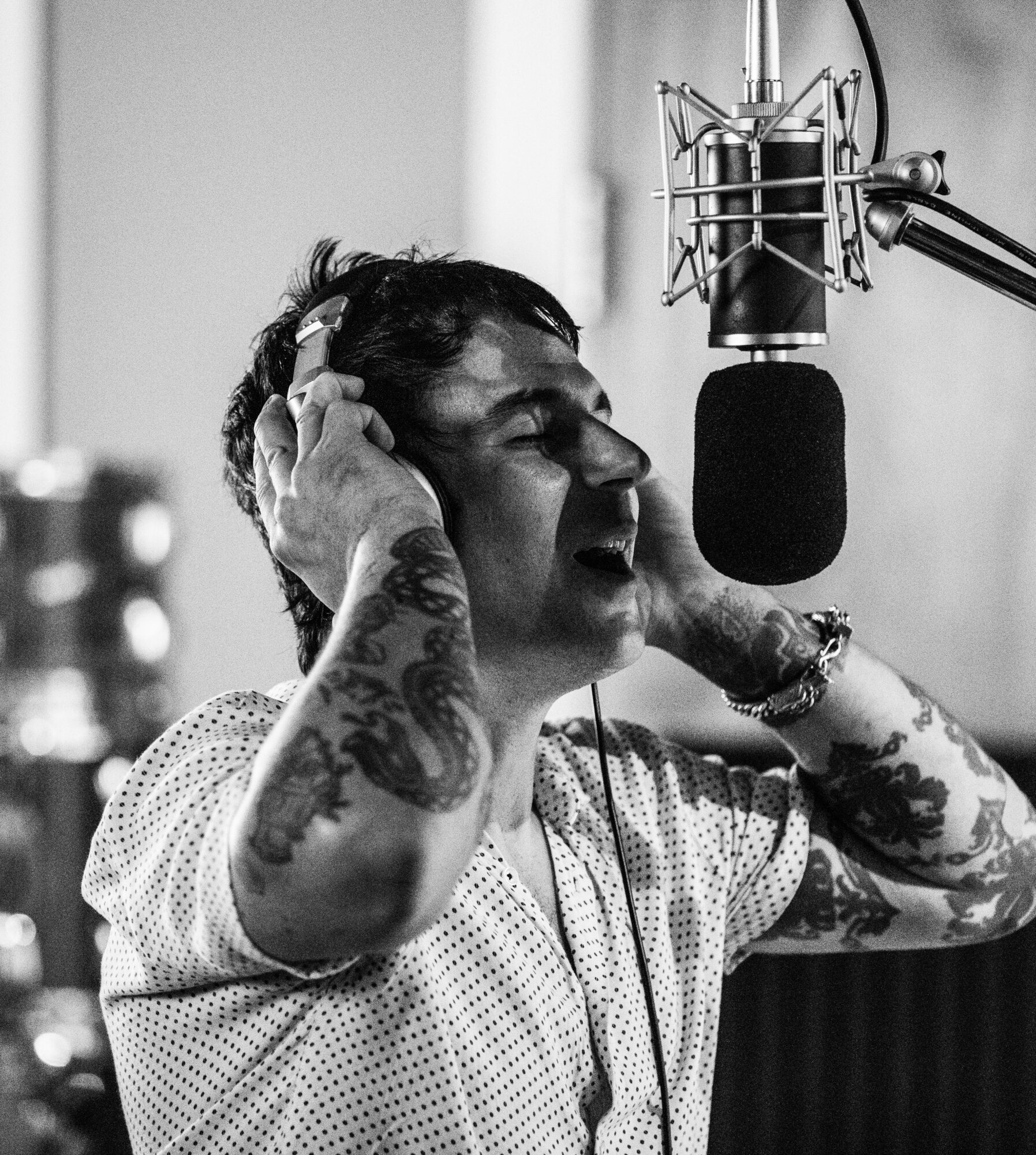 Person singing into a microphone with eyes closed, wearing a patterned shirt, in a recording studio.