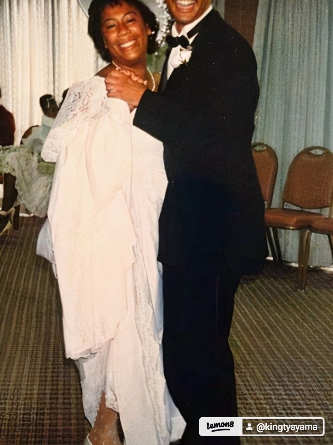 Two people smiling and hugging at a formal event, one in a white dress and the other in a black tuxedo.