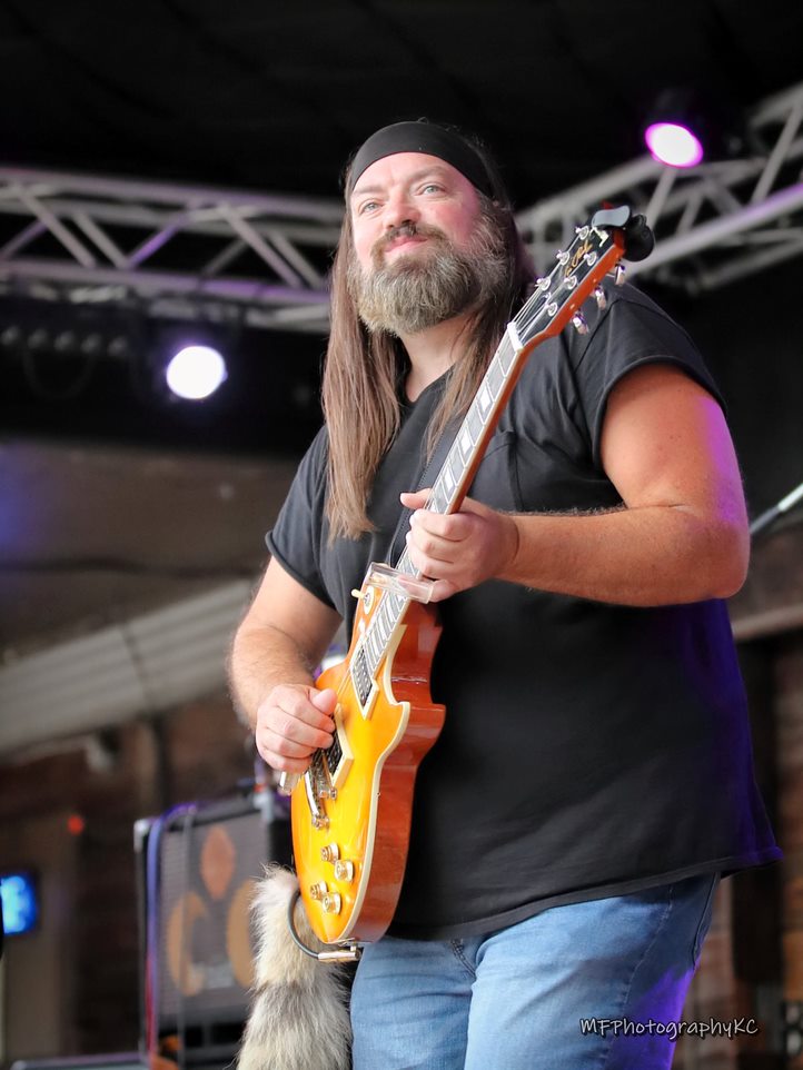 Man with long hair and beard playing yellow electric guitar on stage, smiling, wearing a black headband and black shirt.