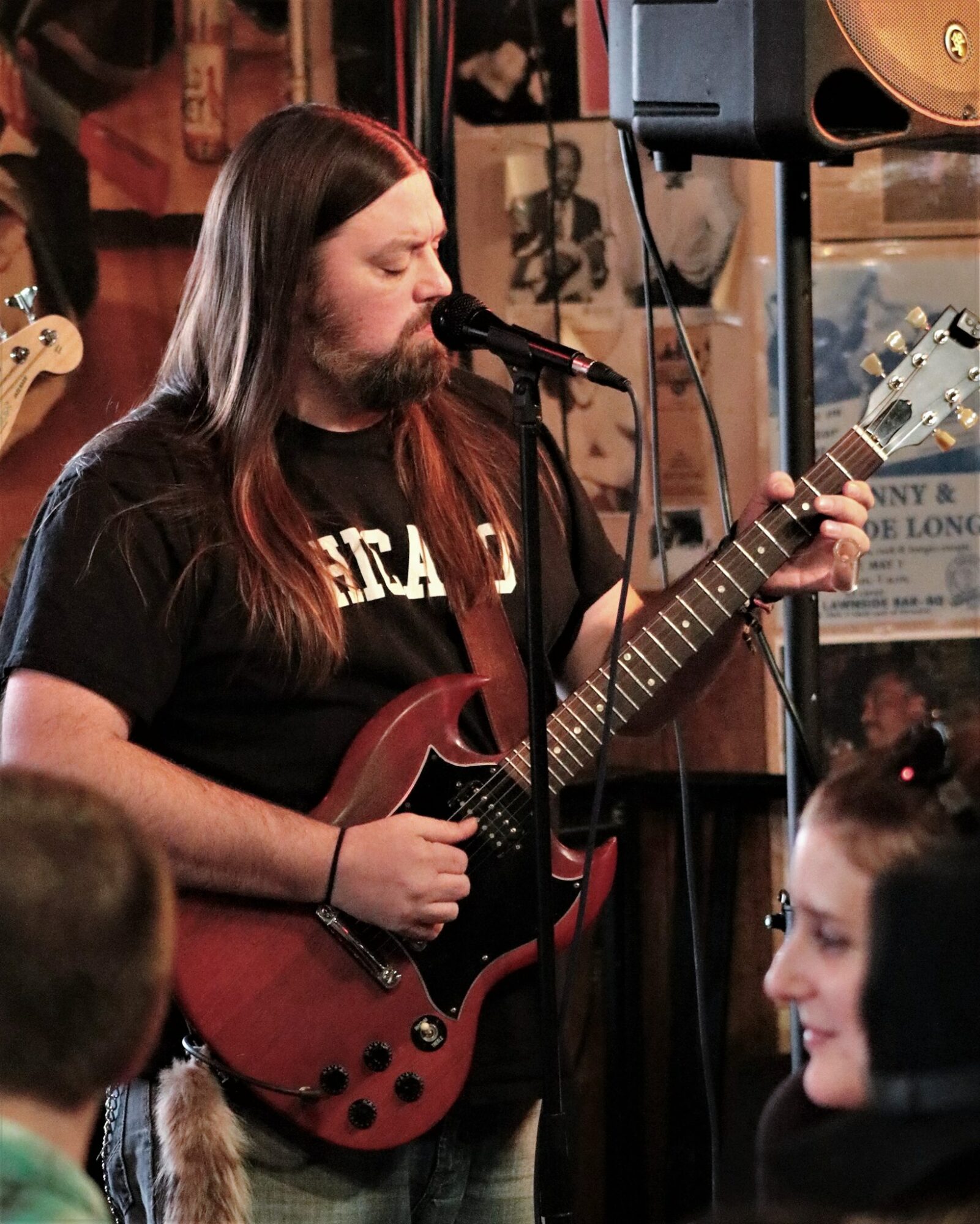 Man with long hair playing electric guitar and singing into microphone in a room with posters and speakers