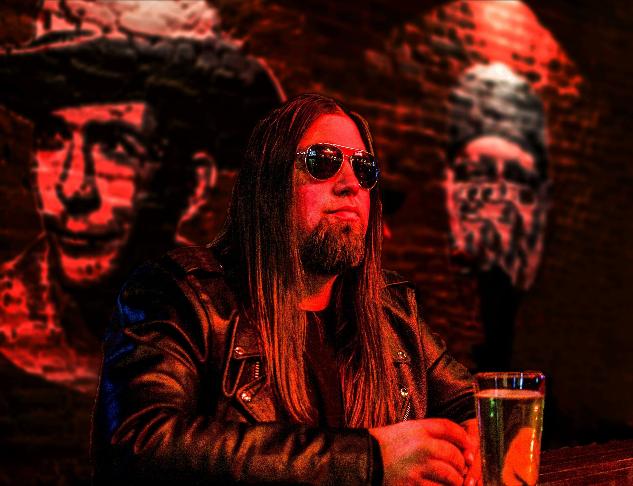 Man with long hair, sunglasses, and a leather jacket sitting at a bar with graffiti-style portraits on the wall behind him.