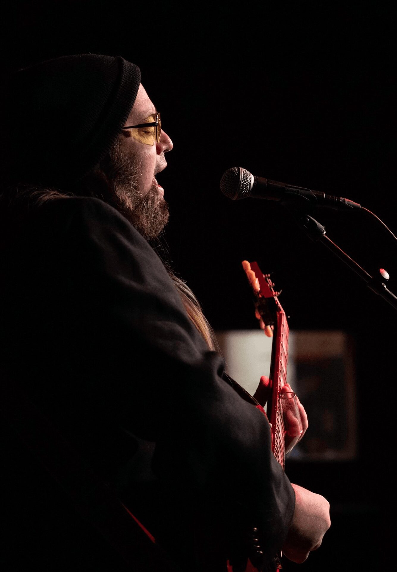 Person with glasses and a beard, wearing a black hat, speaking into a microphone while holding a guitar, against a dark background.