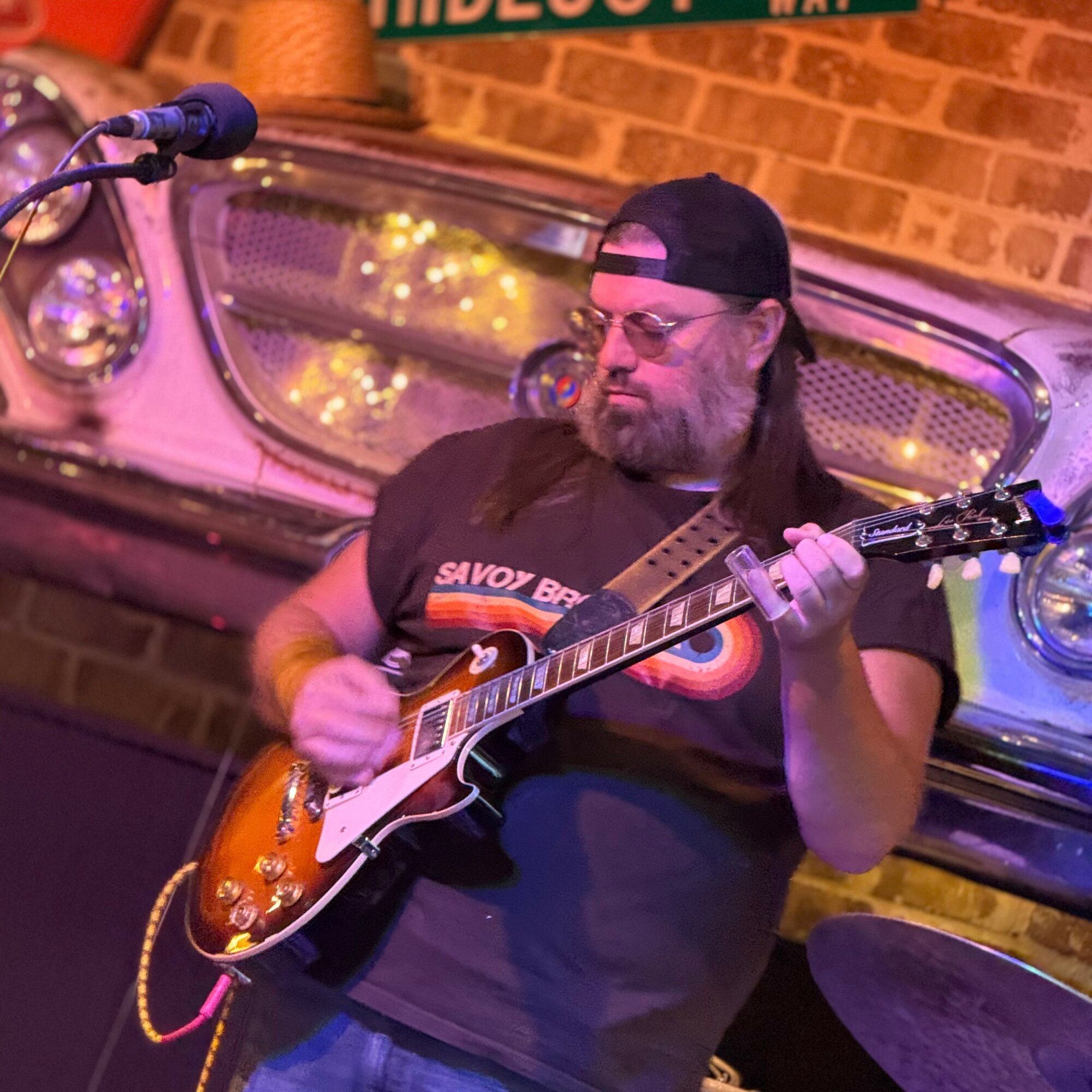 Man playing electric guitar on stage with brick wall background and microphone nearby.