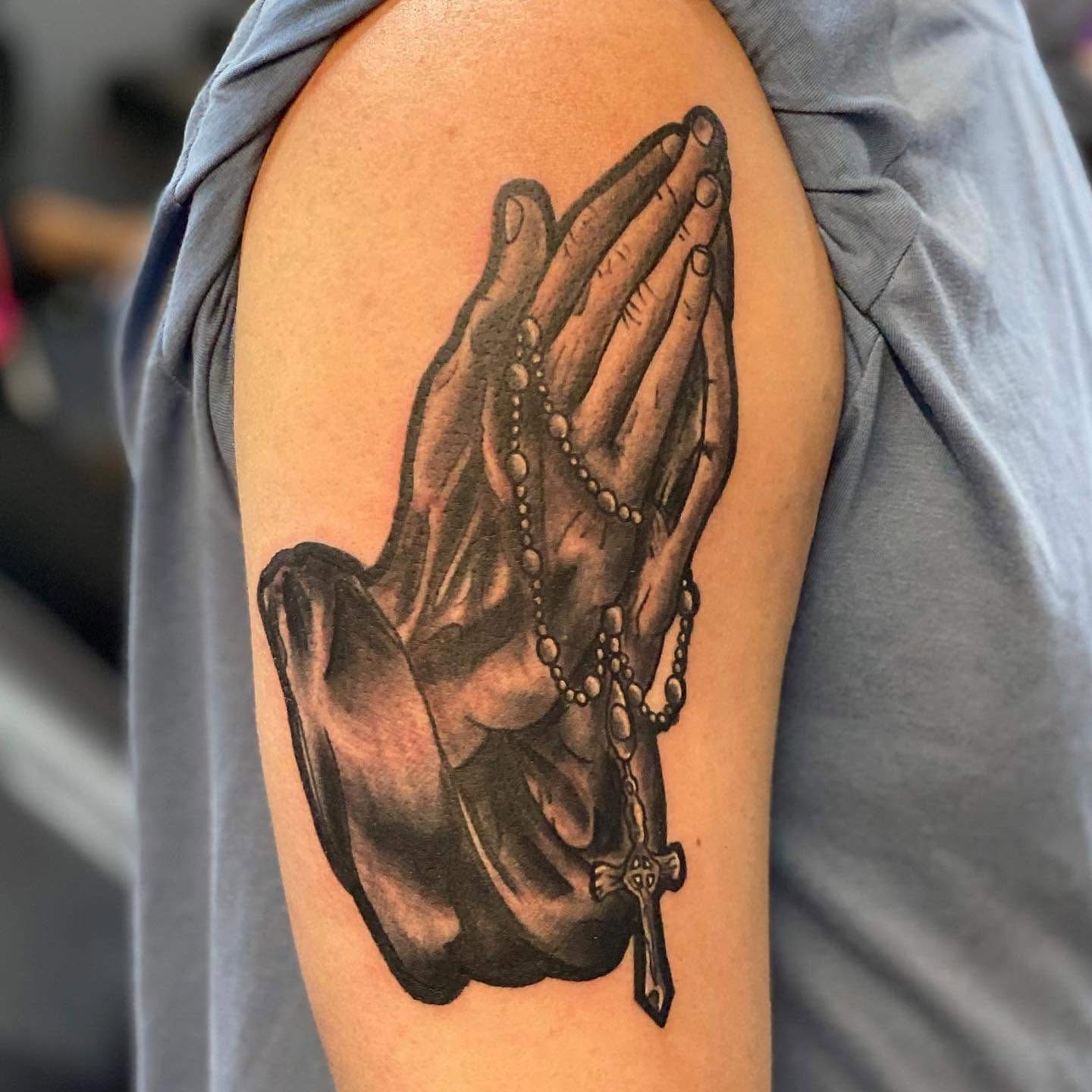 Tattoo of praying hands holding a rosary with a cross on the arm, detailed shading, realistic style.