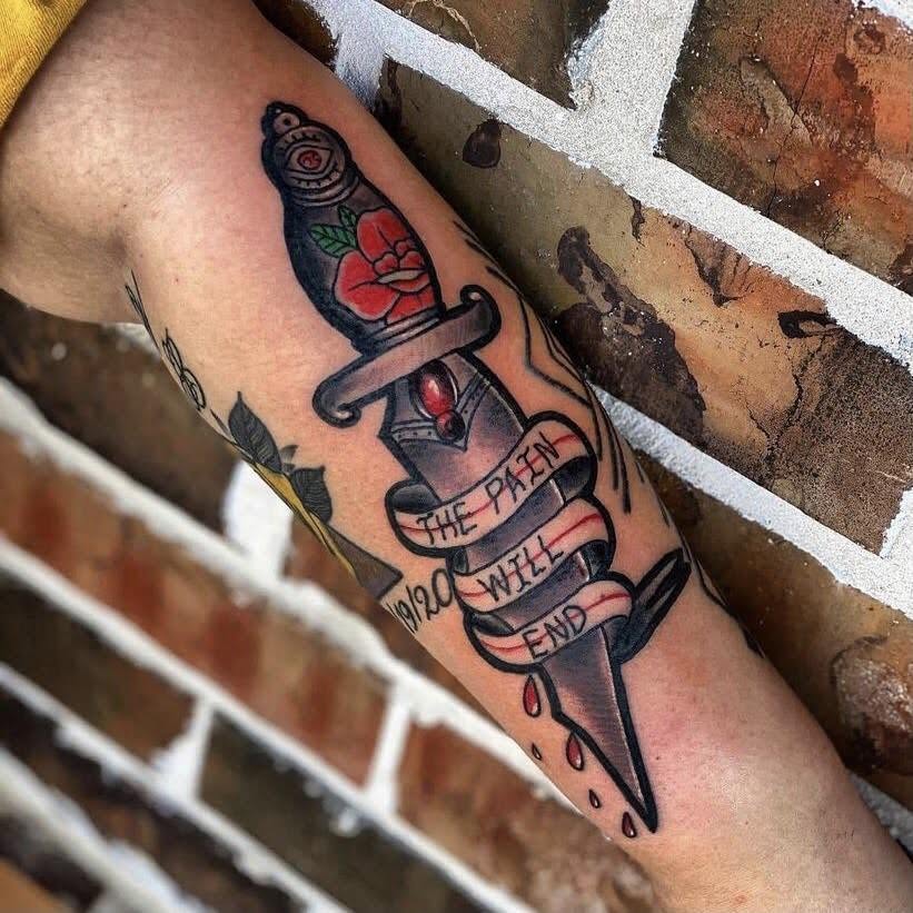 Tattoo of a dagger with a red rose and a banner reading 'The pain will end' on a person's arm.
