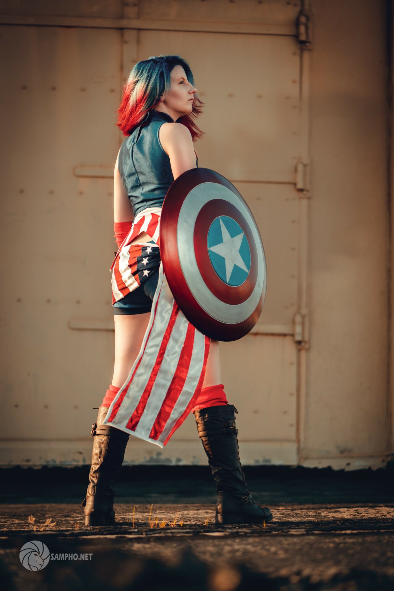 Person dressed as Captain America with shield, standing sideways in front of a beige wall, wearing a costume with stars and stripes.