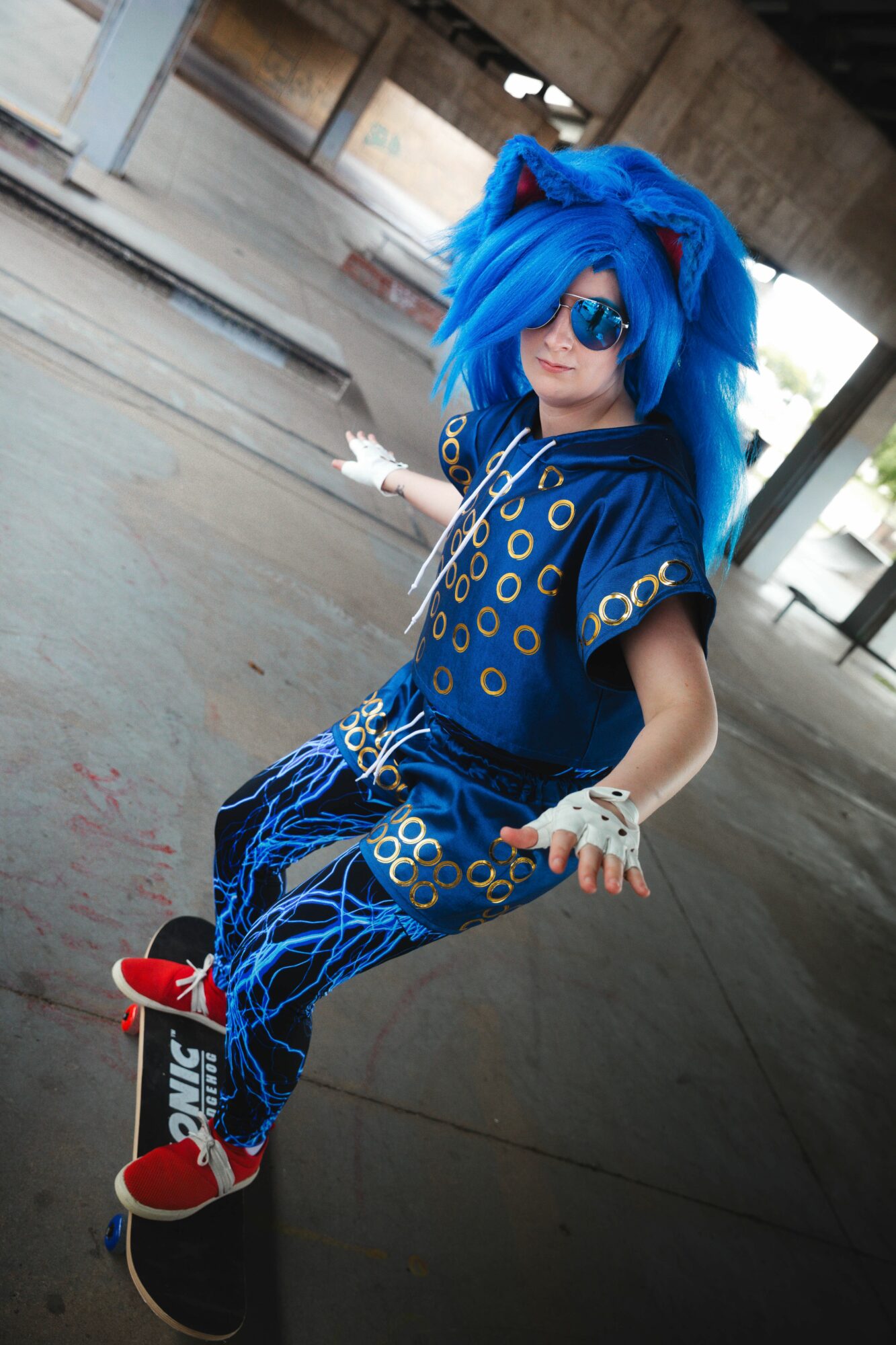 Person with bright blue hair, sunglasses, and a blue outfit with circular patterns, standing under a bridge.