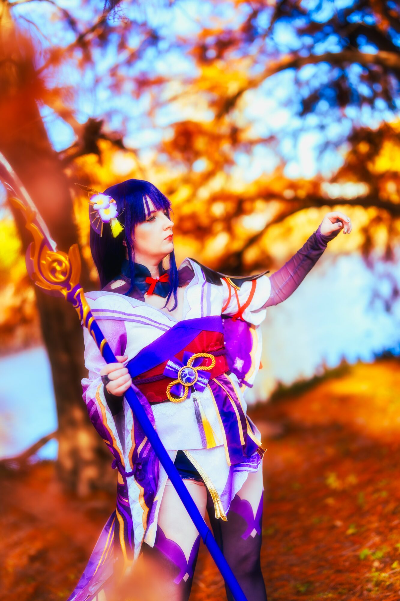 Person in colorful costume with a staff, standing outdoors with autumn trees and sky in background.