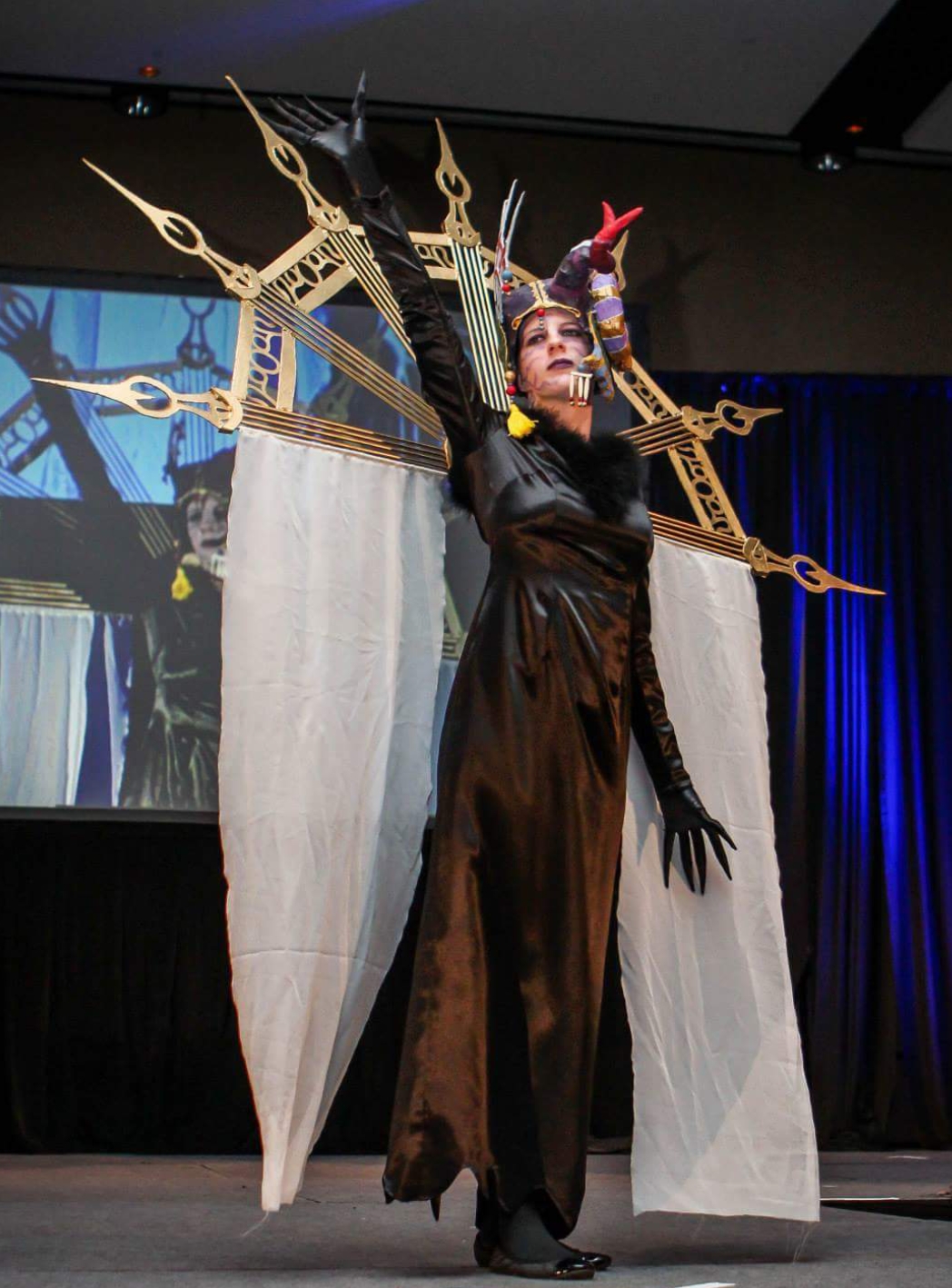 Person in black costume with headgear and makeup, standing in front of a large clock-like structure on stage.