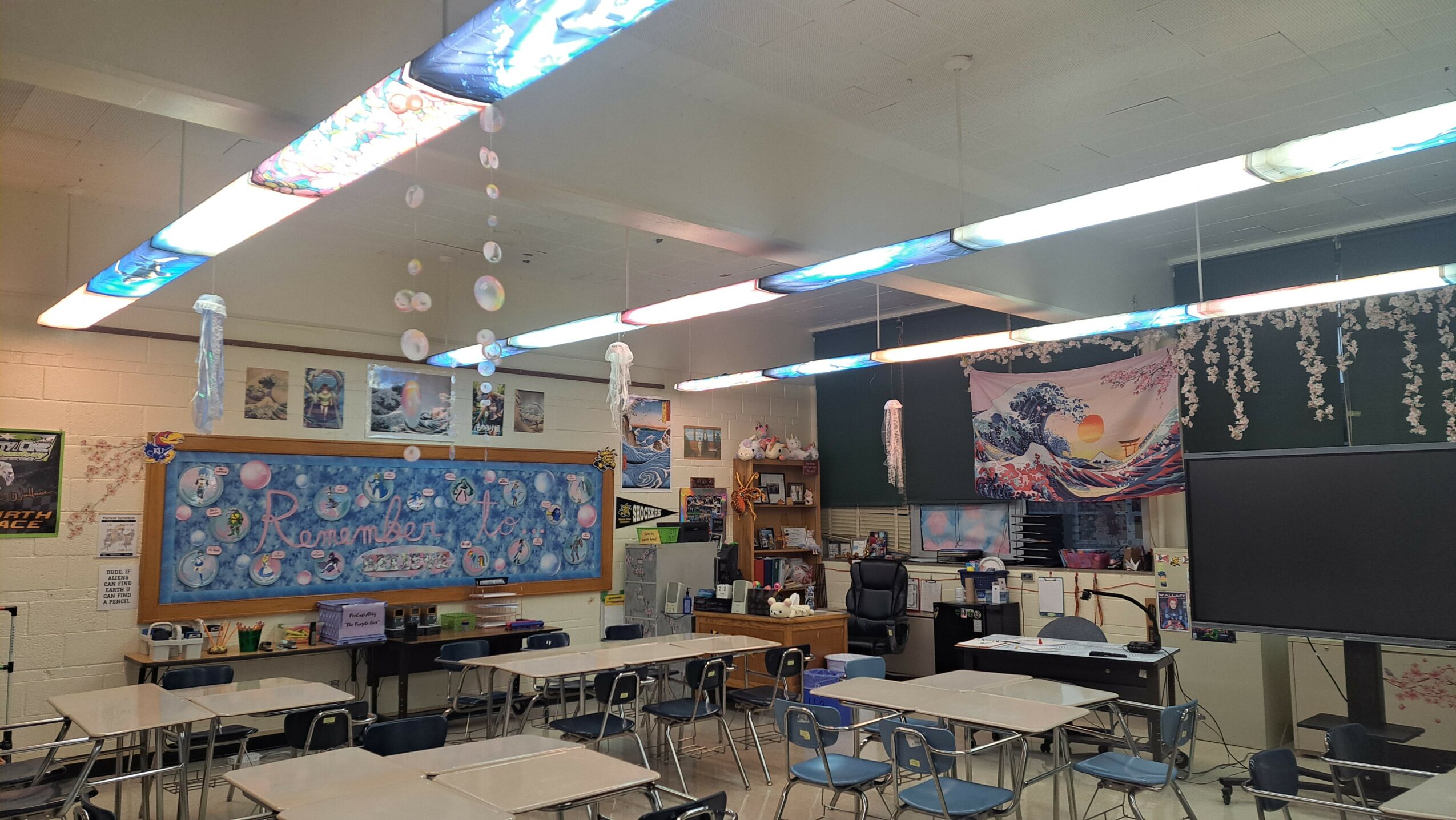 Classroom with desks, chairs, bulletin boards, and a large blackboard, decorated with artwork and educational materials.