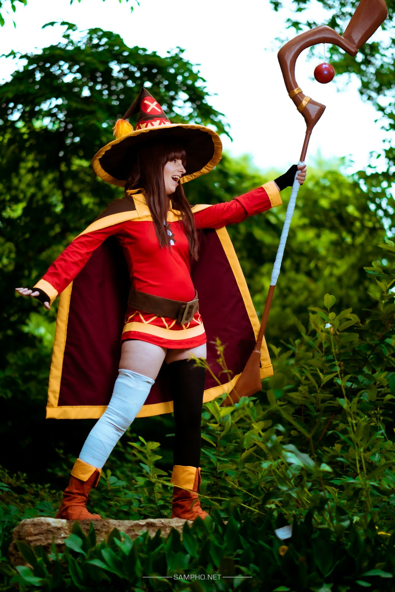 Person in colorful costume with a large hat and cape, holding a staff with a ball, standing outdoors among greenery.