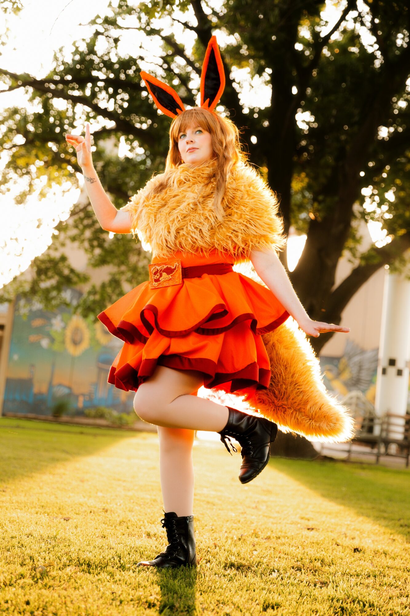 Person dressed as a fox with ears, tail, and furry costume, standing outdoors on grass, one leg raised, arms outstretched.