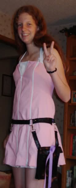 Young woman with curly hair smiling and making a peace sign, wearing a pink dress with a black belt.
