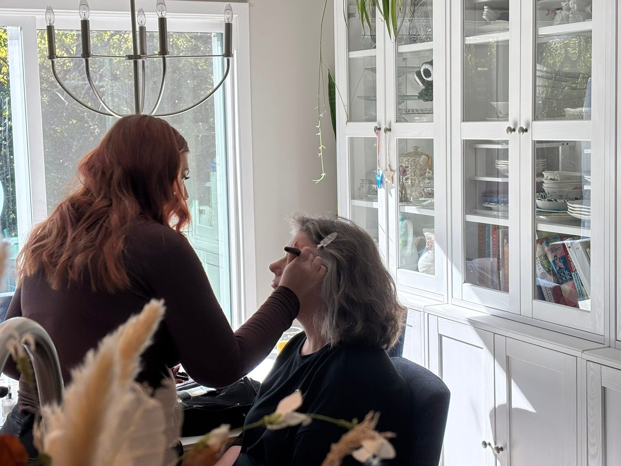 Two women in a bright room, one applying makeup to the other, with glass cabinets and a window in the background.