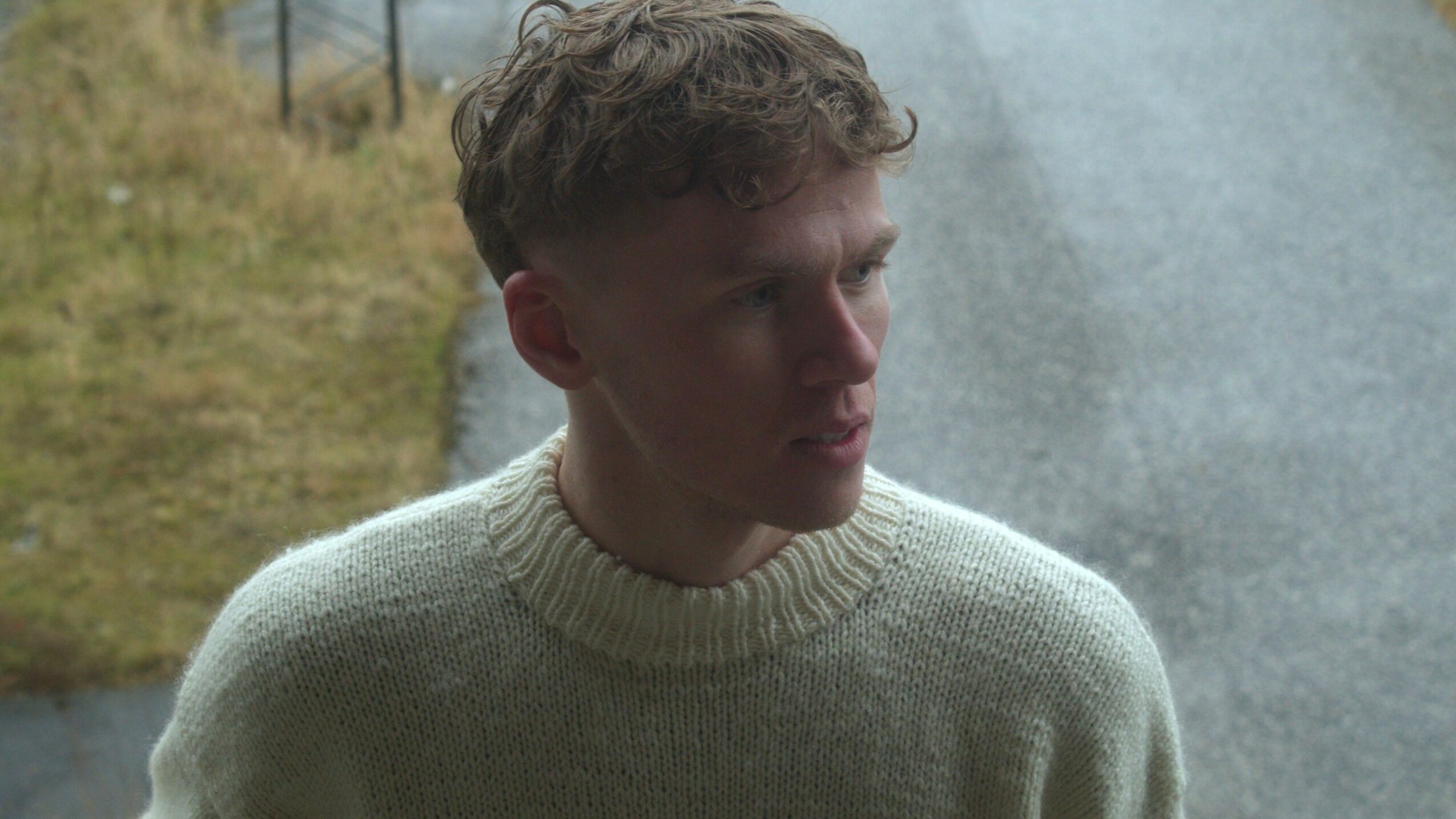 Young man with curly hair wearing a light-colored sweater, looking to the side outdoors.
