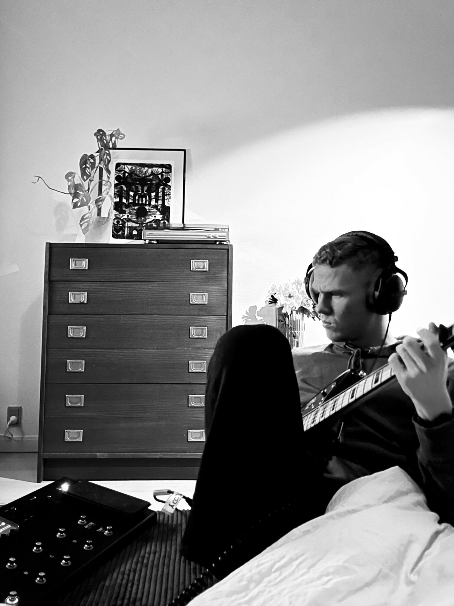 Person sitting on bed playing guitar, wearing headphones, with a dresser and framed photos in the background.