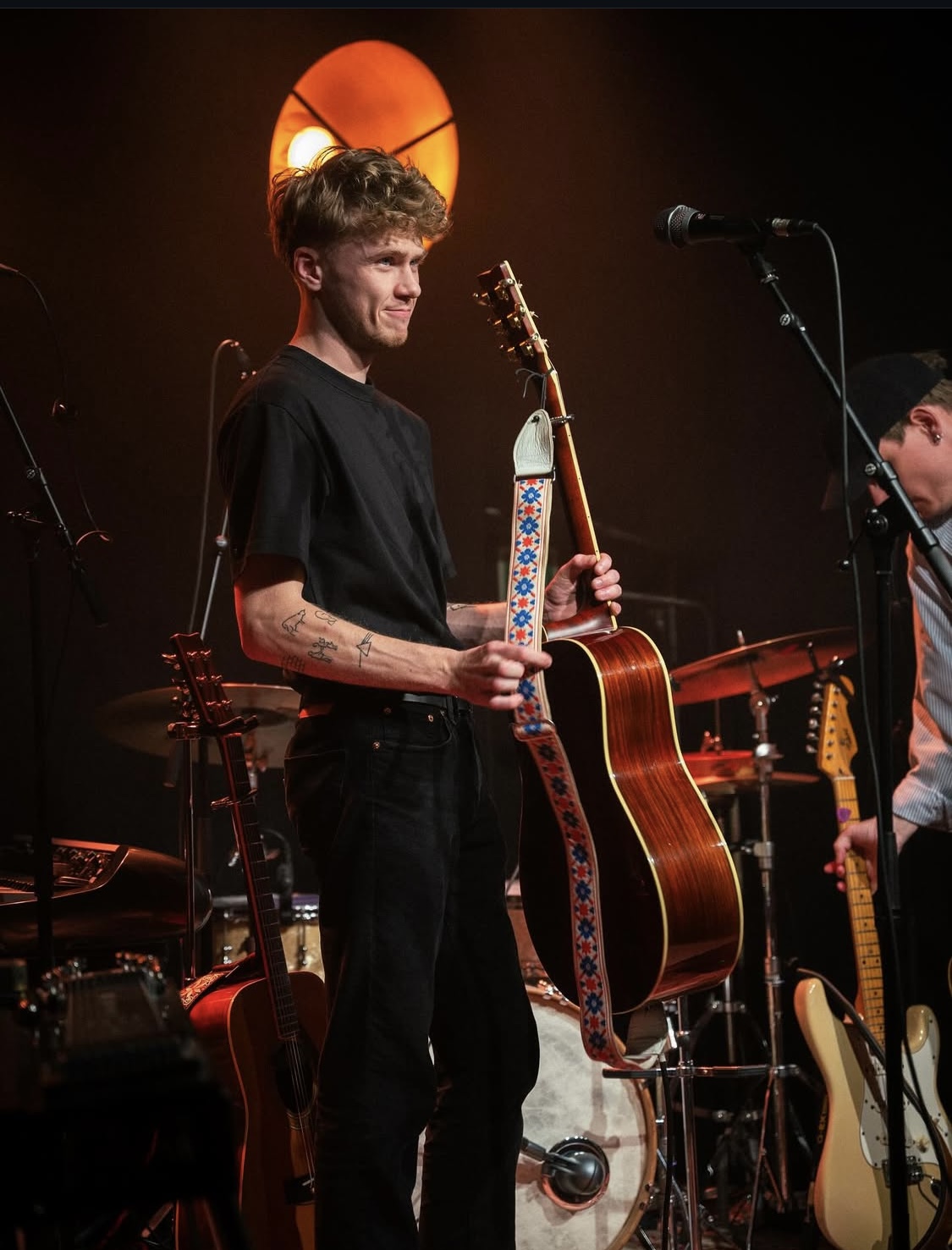 Young man with short hair holding a guitar strap, standing on stage with musical instruments and microphone, smiling.