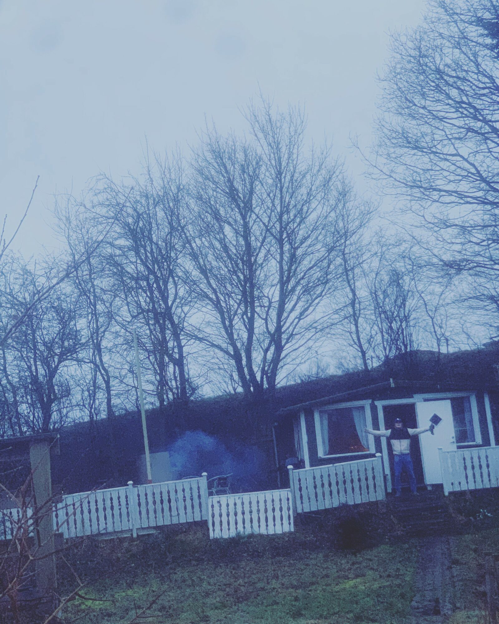 A person stands on a porch with a white railing, trees without leaves are in the background, sky is overcast.