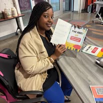 Woman sitting at a table holding a paper, with books and a backpack nearby, in an indoor setting.