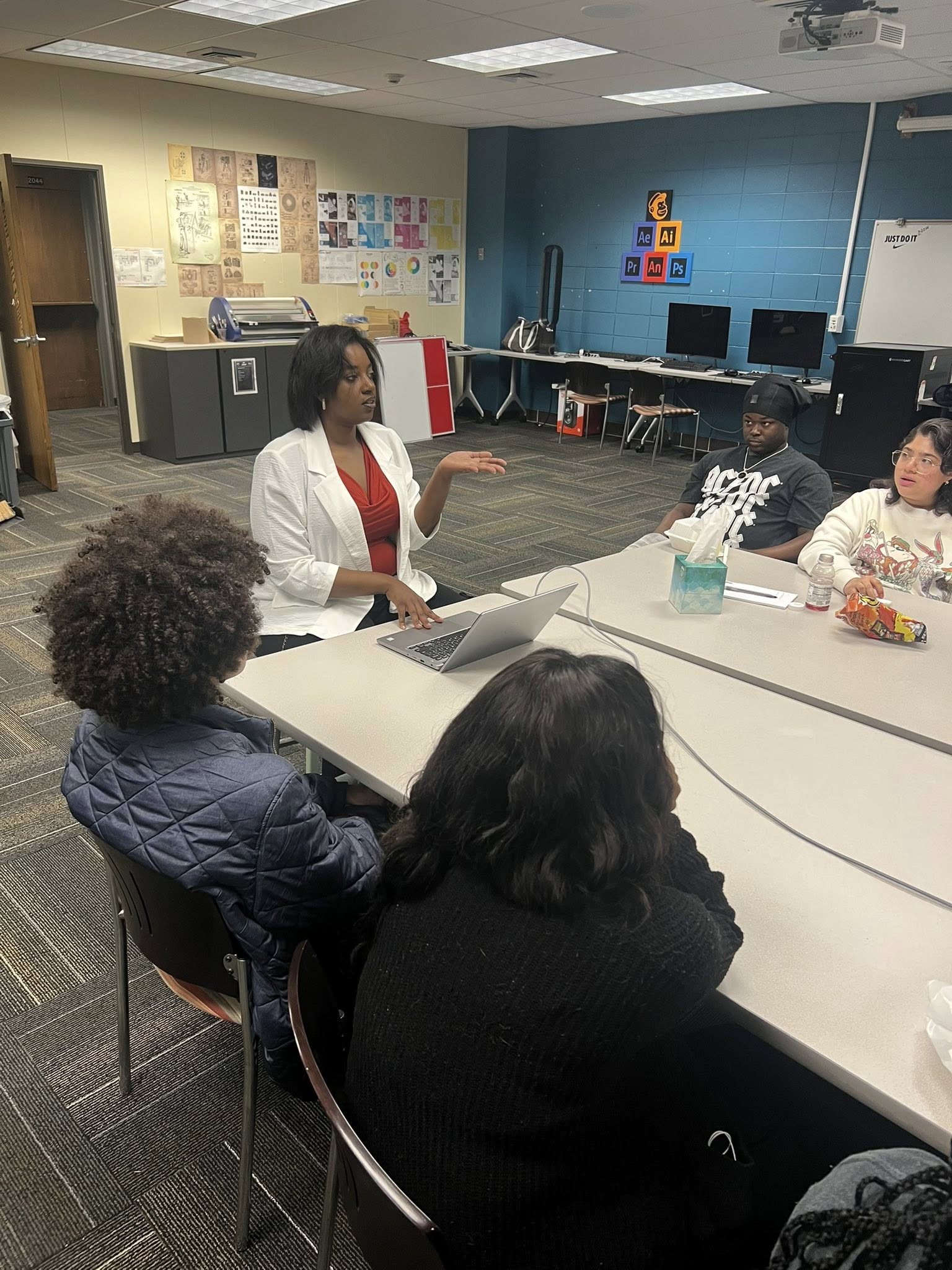 A woman standing and speaking to a group of four seated people in a classroom with computers and colorful posters.