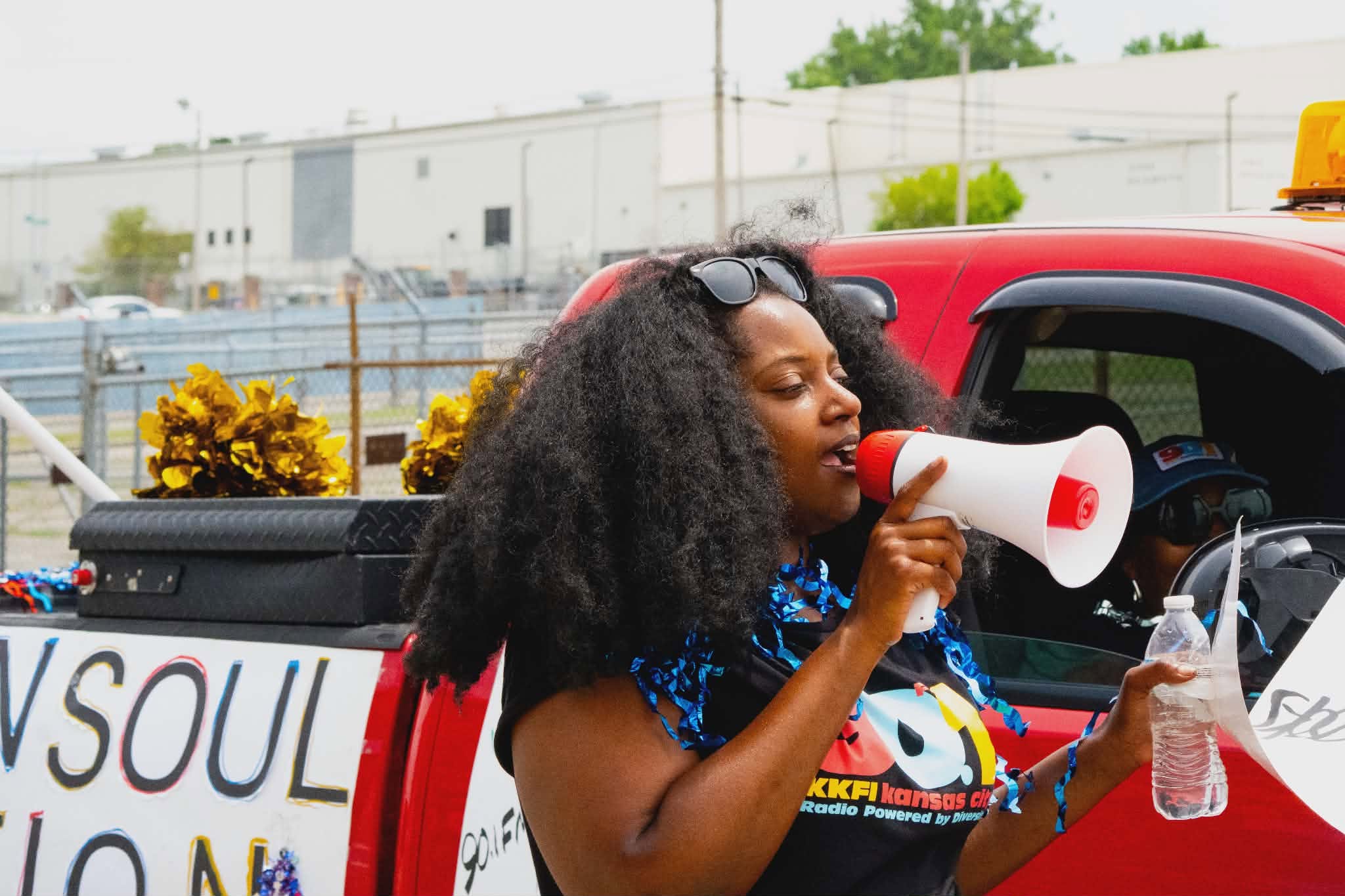 Woman with curly hair using a megaphone and holding a water bottle near a red vehicle, outdoors with yellow flowers and buildings in background.