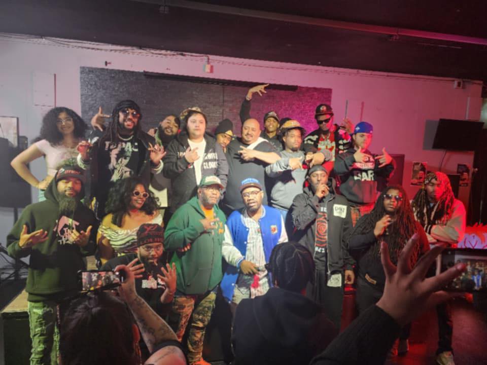 Group of people on stage, some holding microphones, in a dimly lit room with pink and purple lighting, celebrating.
