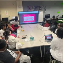 Group of children and adults seated around a large table watching a presentation on a screen in a classroom.