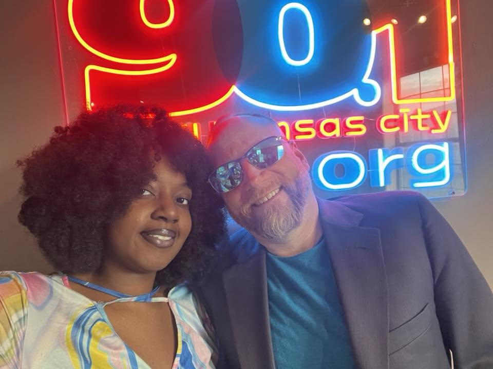 Two people smiling in front of a neon sign with red, blue, and yellow lights, reading 'Kansas City' and 'org'.