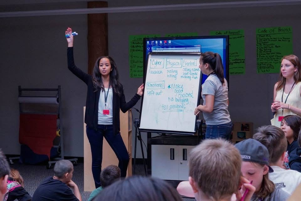 A woman stands in front of a group, gesturing with her right hand, with three women and an audience of seated people behind her, in a conference room with green posters on the wall.
