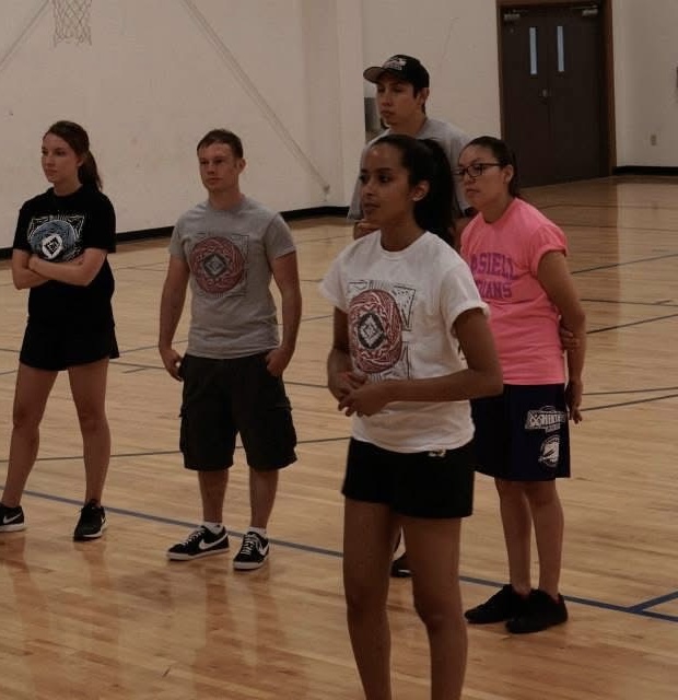 Group of five children standing on a gymnasium floor, facing forward, with a door in the background.