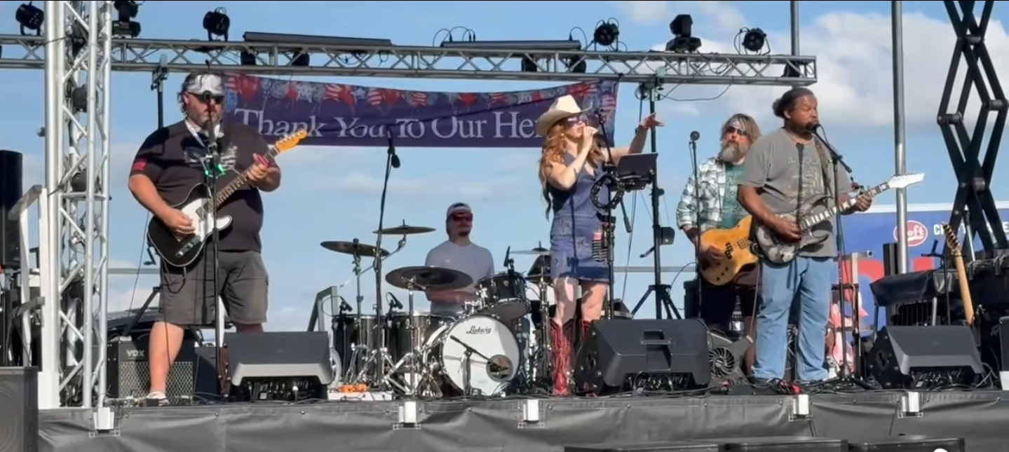 Band performing on stage with five members, including a singer, guitarist, drummer, and others, outdoors during daytime.