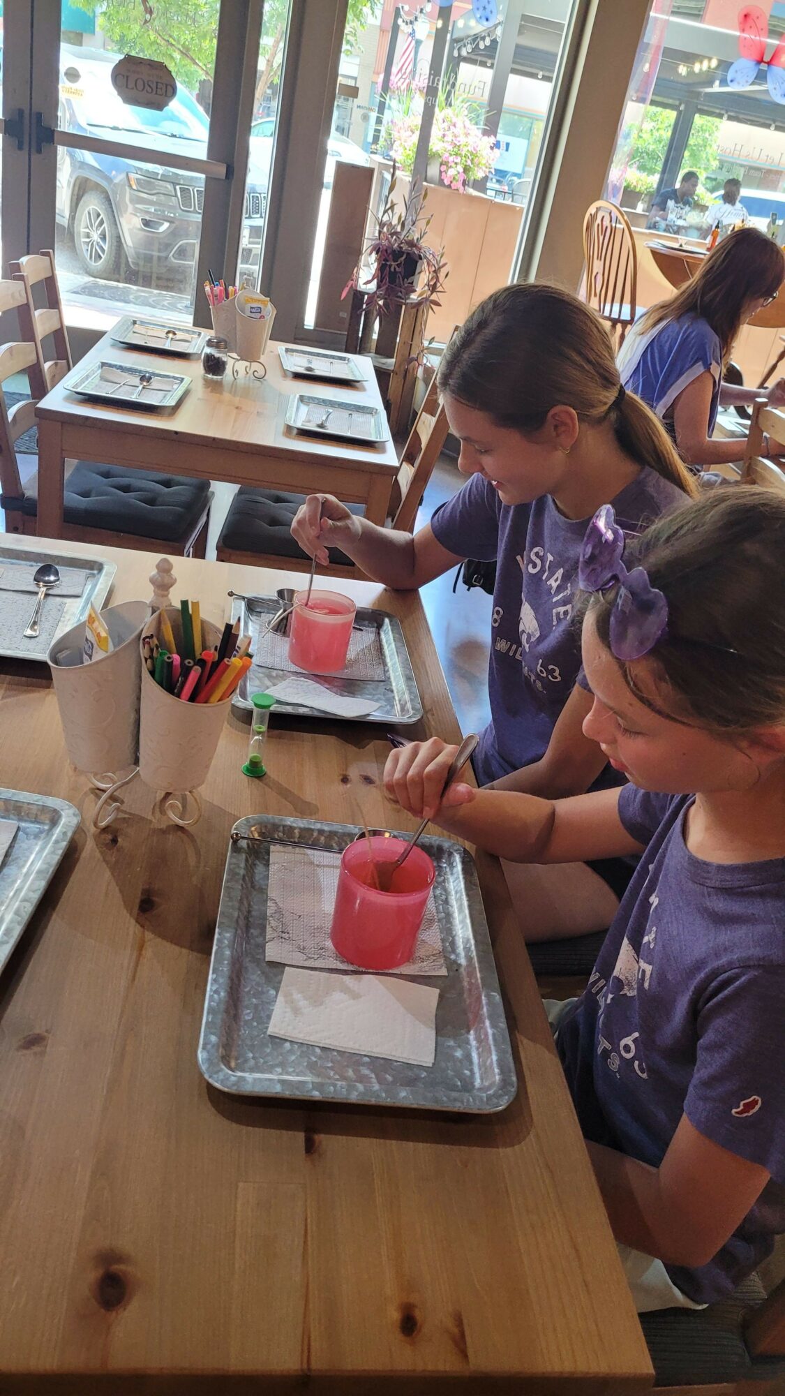 Two children are engaged in a craft activity at a wooden table, with art supplies and a red cup on a tray.
