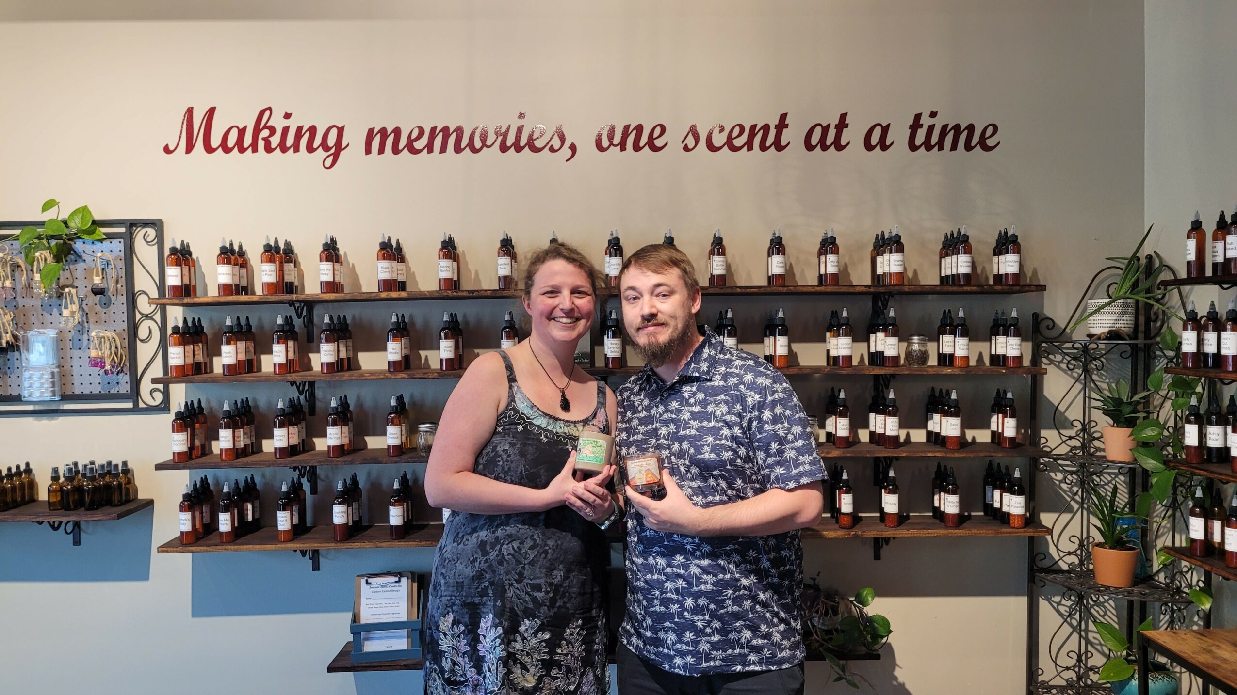Two people holding wine glasses in front of shelves with small bottles, smiling, with a sign above reading 'Making memories, one scent at a time'.