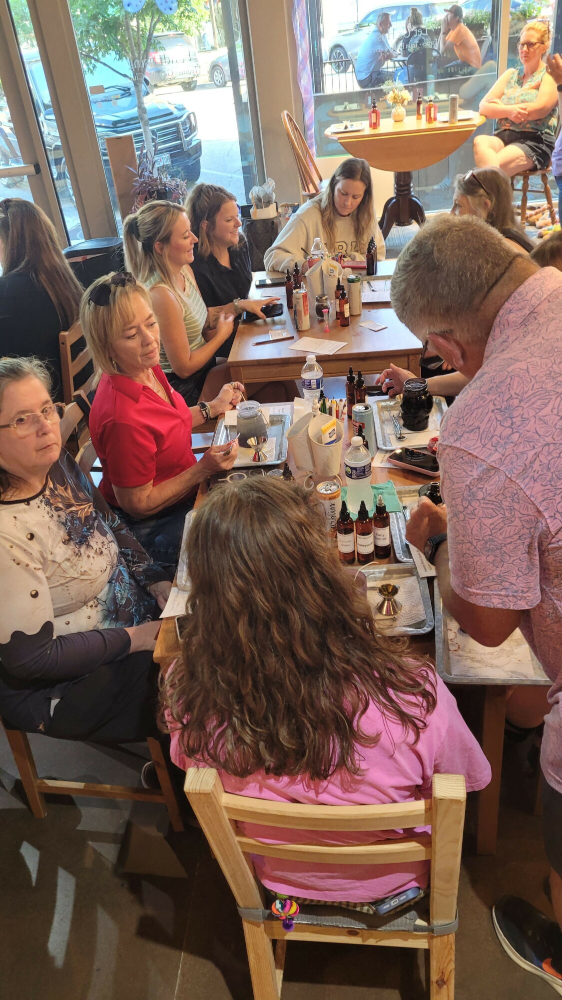 People sitting at tables in a busy restaurant or cafe, engaging in conversation and dining, with large windows in the background.