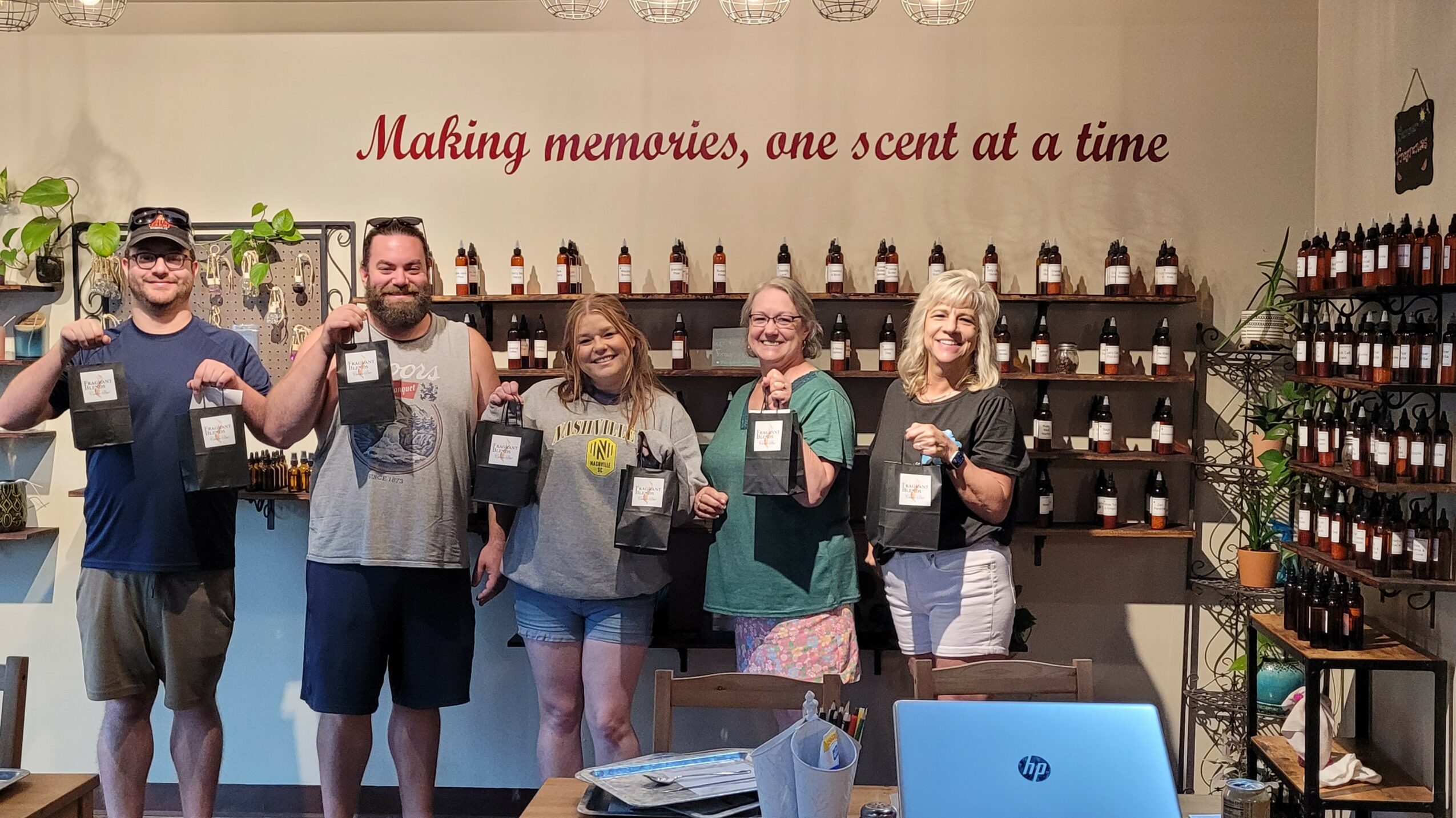Five people stand in a store holding bottles, smiling, with shelves of bottles behind them, and a sign above reading 'Making memories, one scent at a time'.