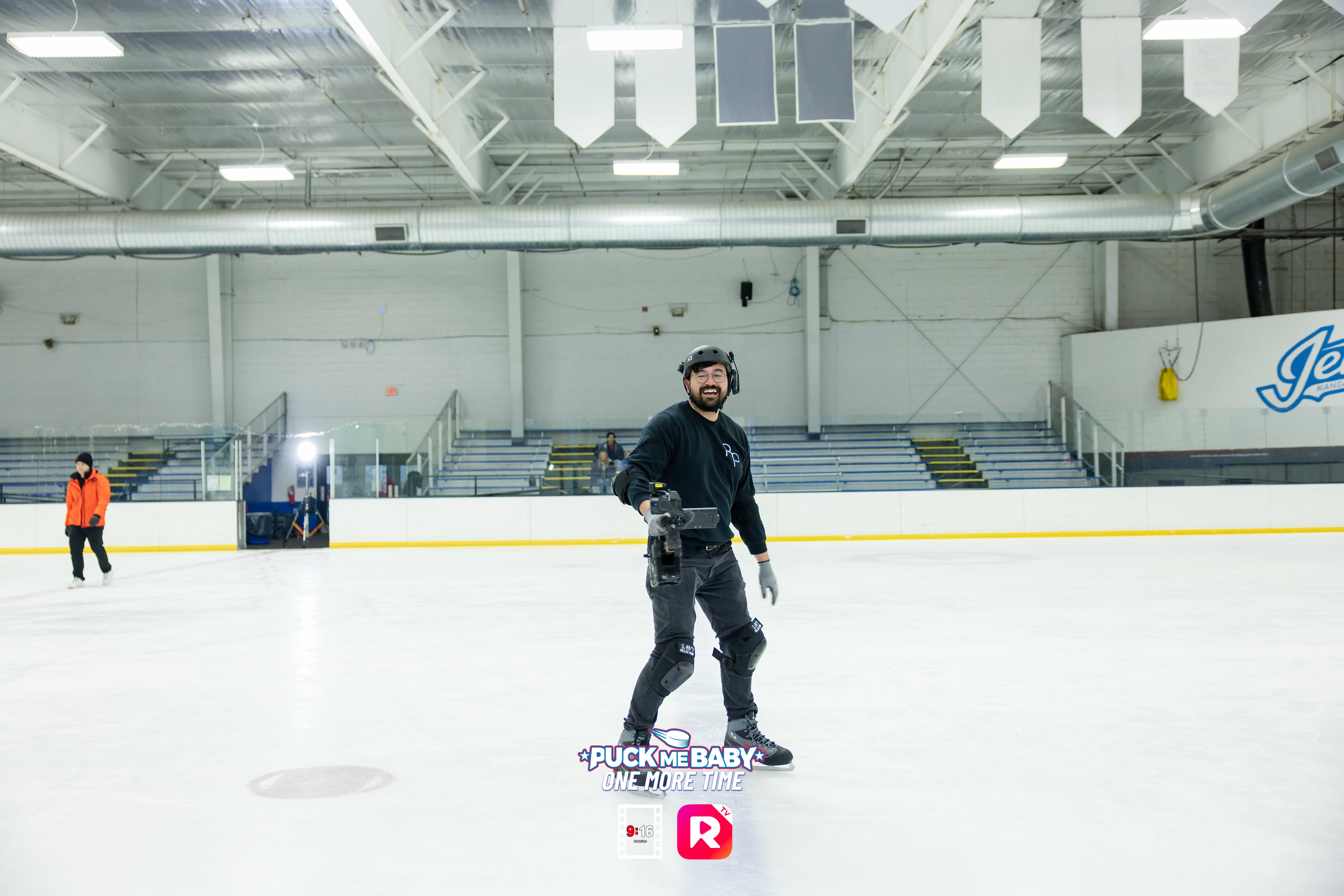 Person standing on ice rink holding hockey stick, wearing helmet and gear, with empty seating and rink in background.