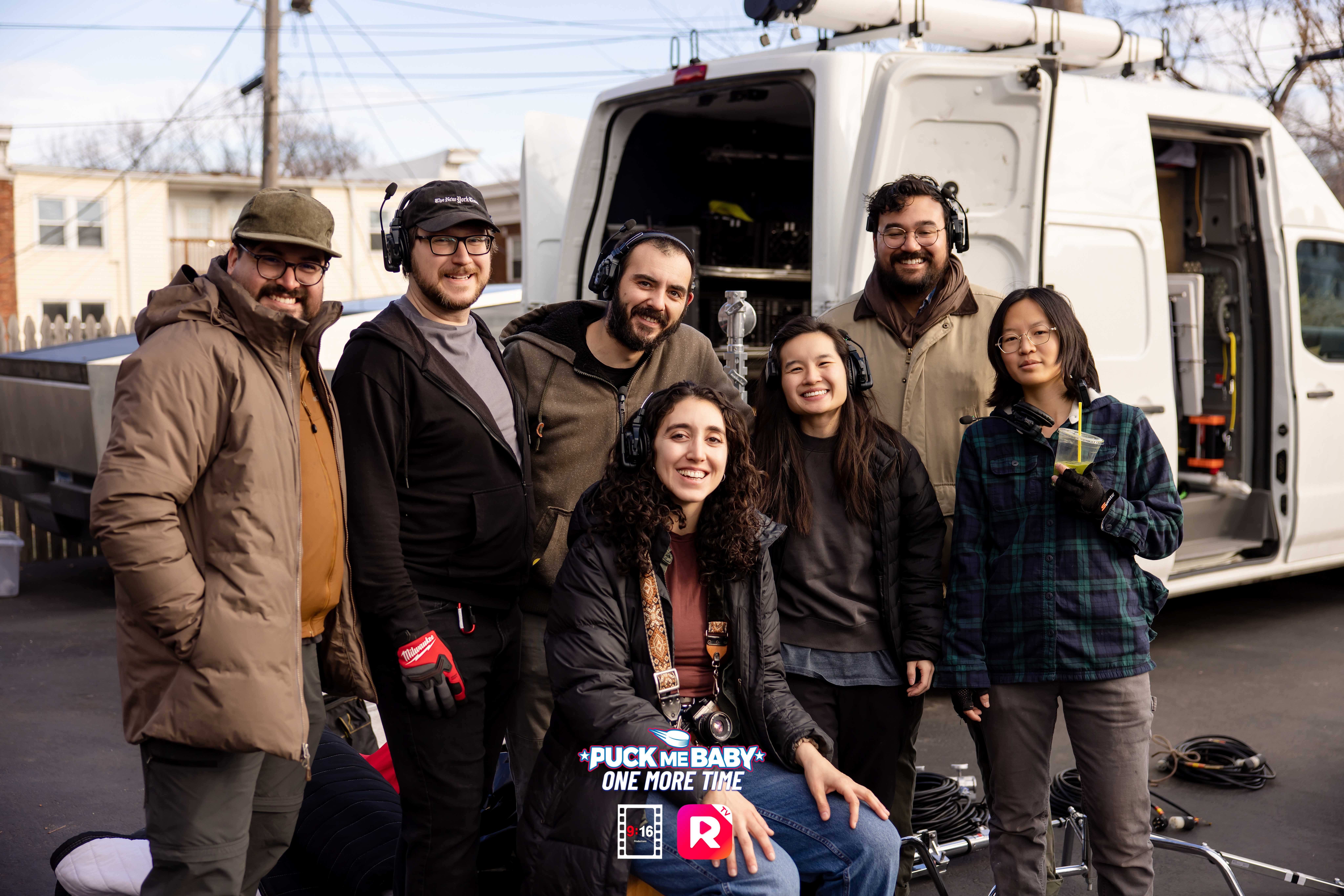 Group of seven people standing outdoors near a white van, smiling, with buildings and power lines in background.