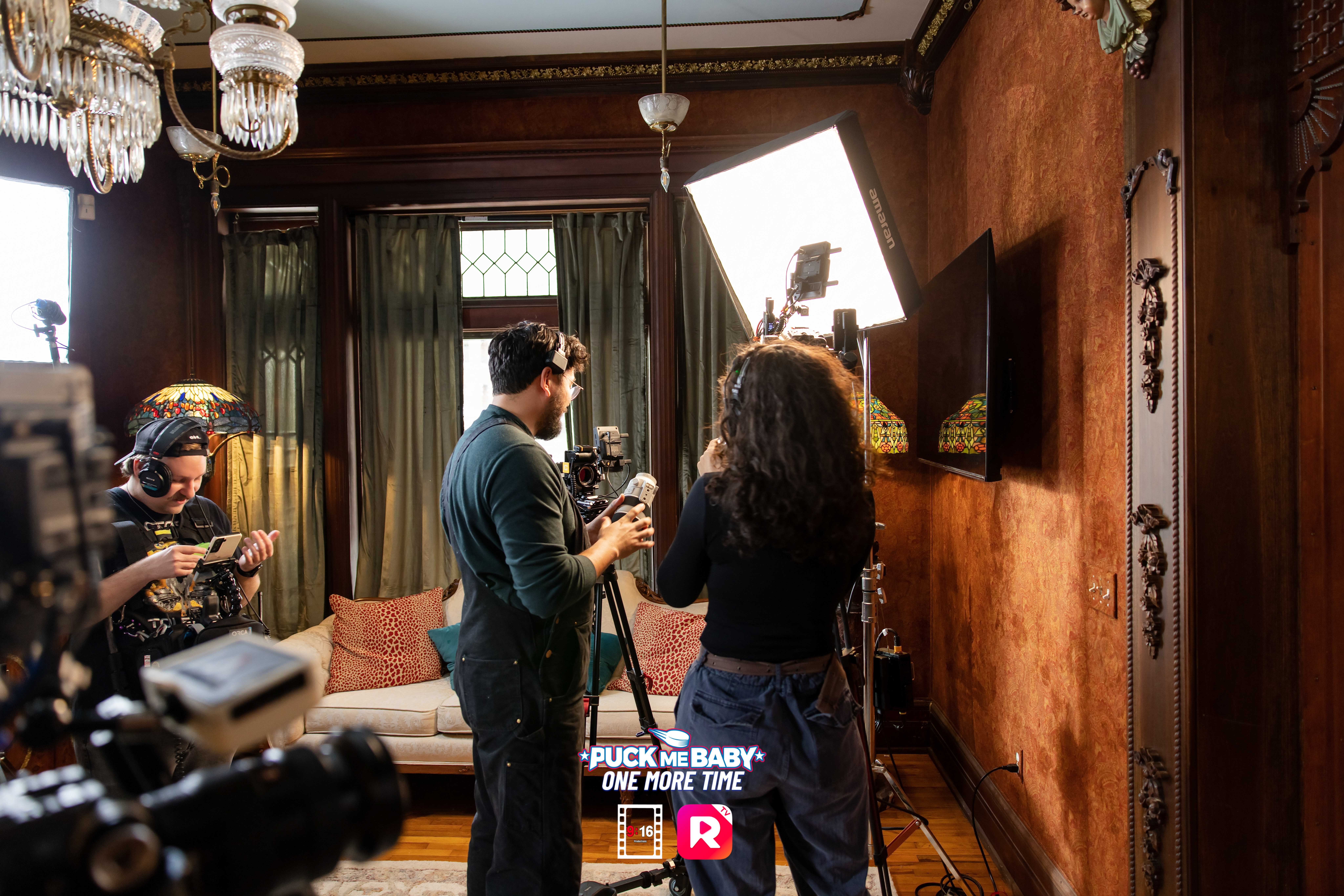 People in a room with wooden walls and a large window, filming a woman with curly hair, a man, and cameras.