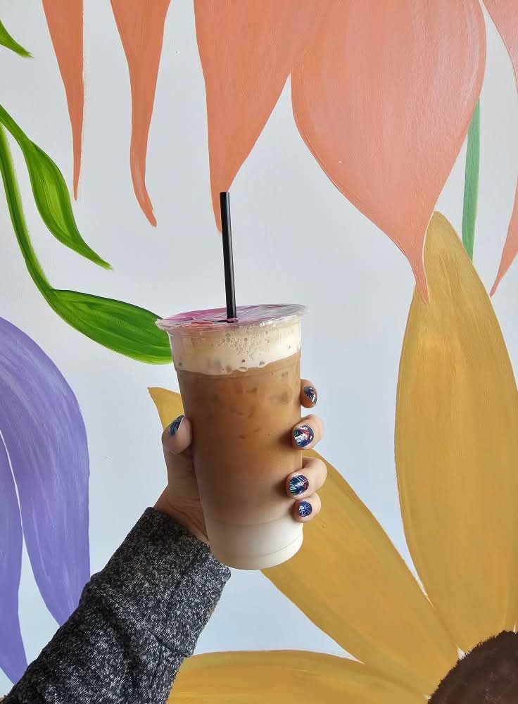 Person holding a tall iced coffee with a straw in front of a colorful floral mural, hand in a gray sleeve.