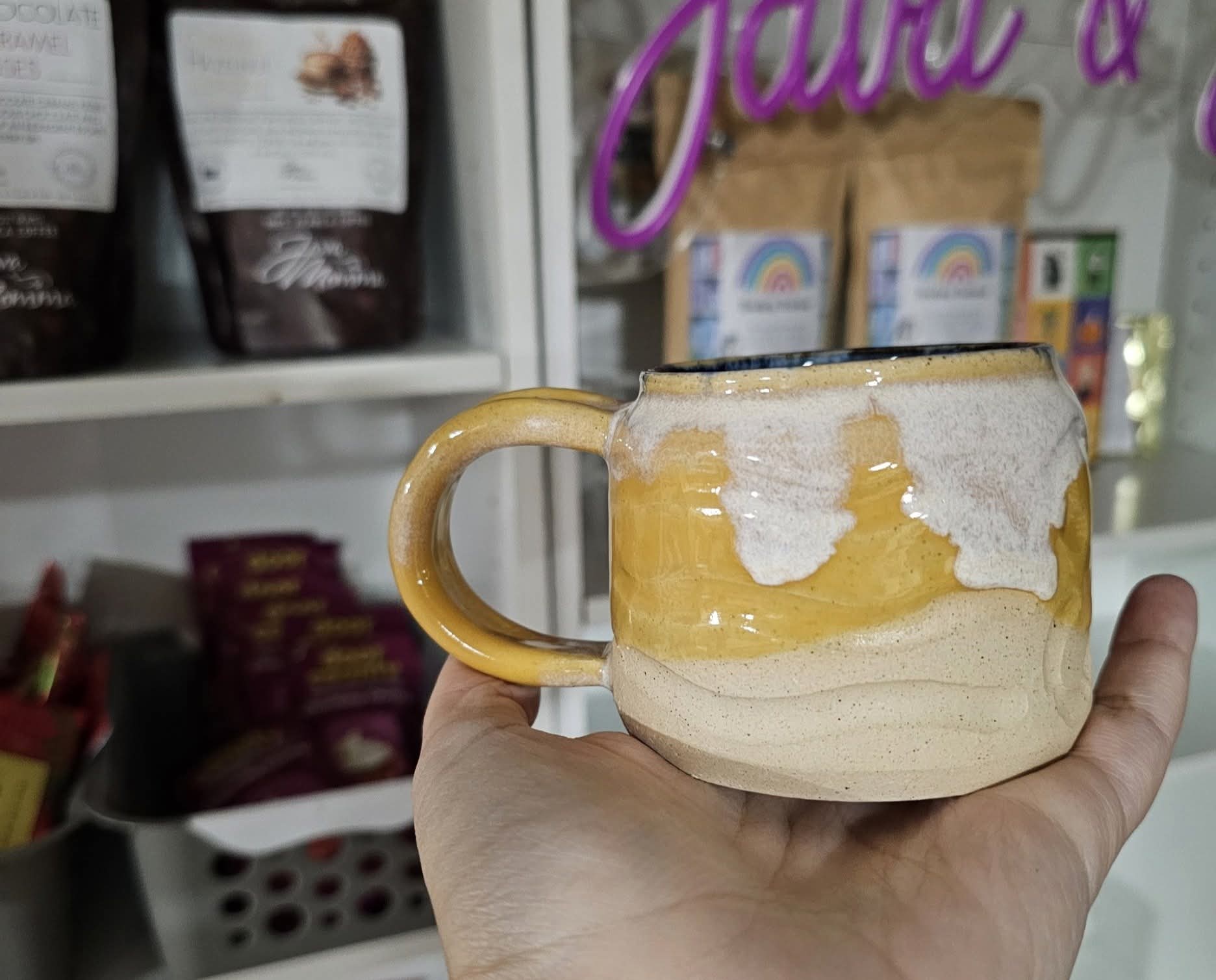 Hand holding a large ceramic mug with a yellow handle, filled with a frothy beverage, in front of store shelves.