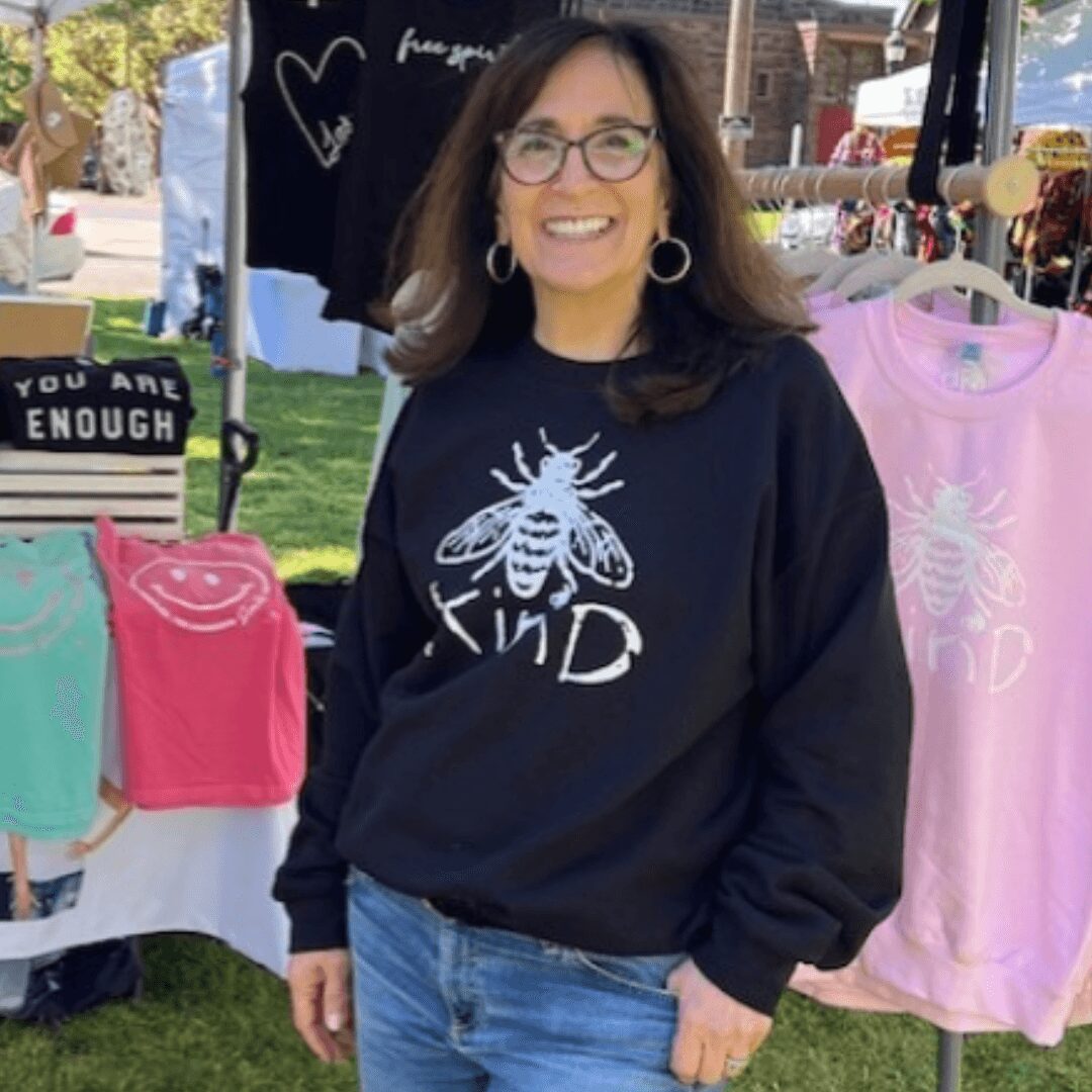 Woman with glasses and hoop earrings smiling outdoors, wearing a black sweatshirt with a bee and the word 'KIND', standing near clothing displays.
