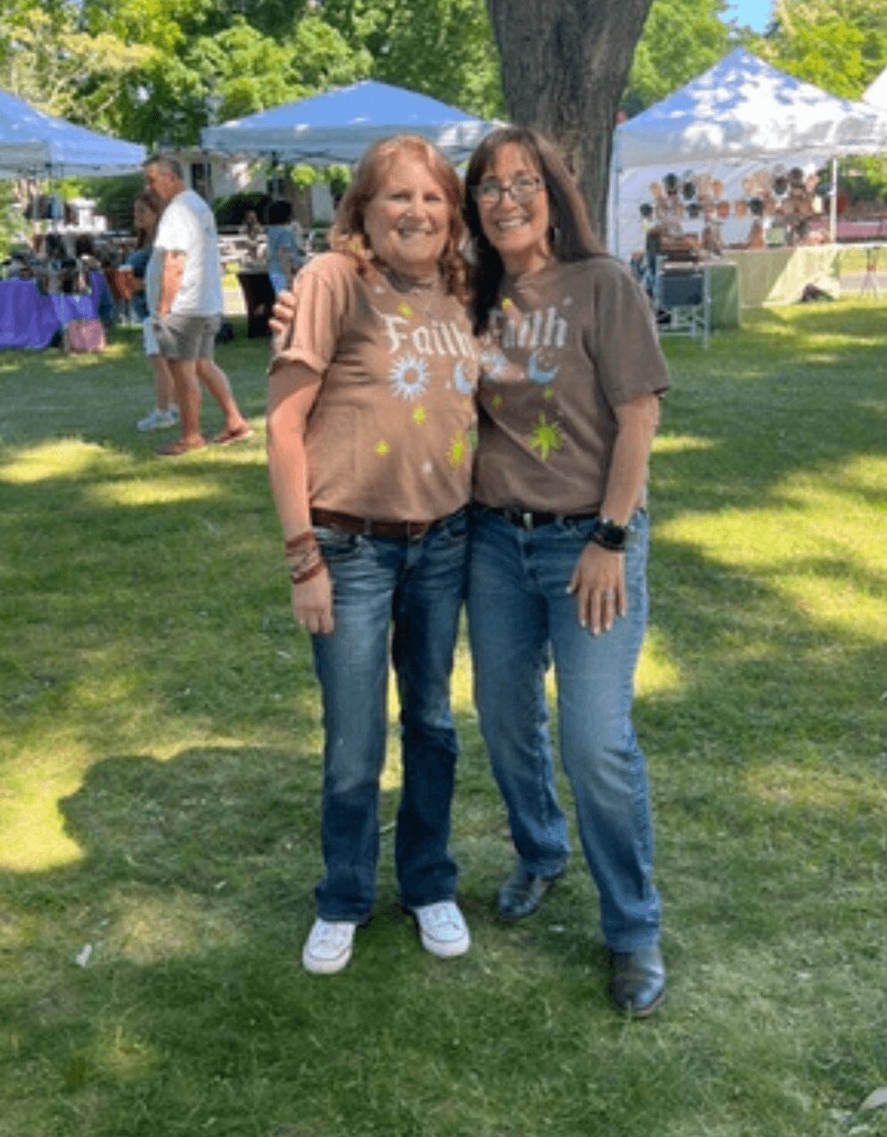Two women stand outdoors on grass, smiling, with tents and trees in the background.