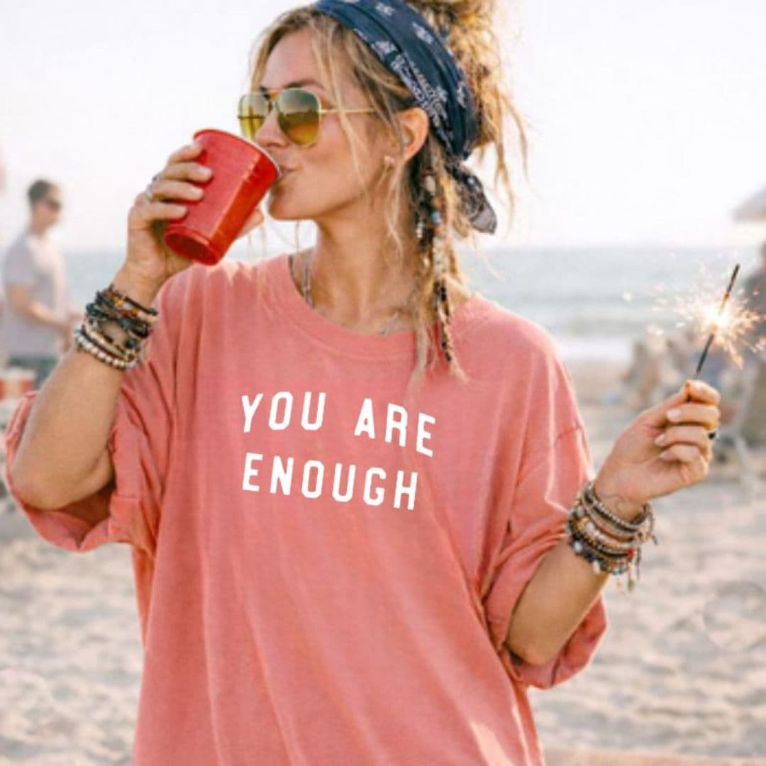 Woman on beach wearing sunglasses and a pink shirt with text, holding a red cup and a sparkler.