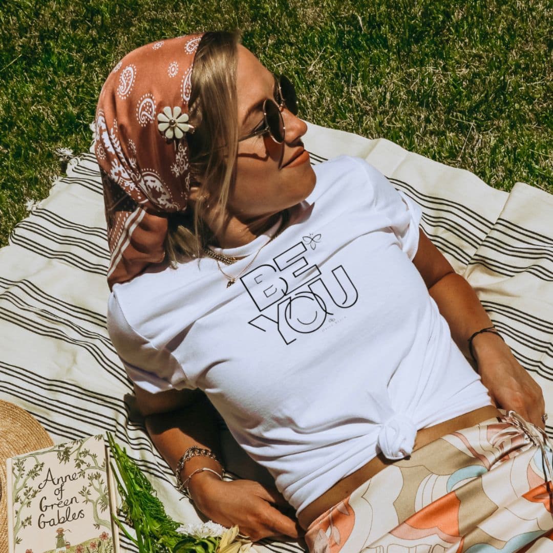 Person lying on a striped blanket outdoors, wearing sunglasses, a white T-shirt, and a patterned headscarf, relaxing in sunlight.