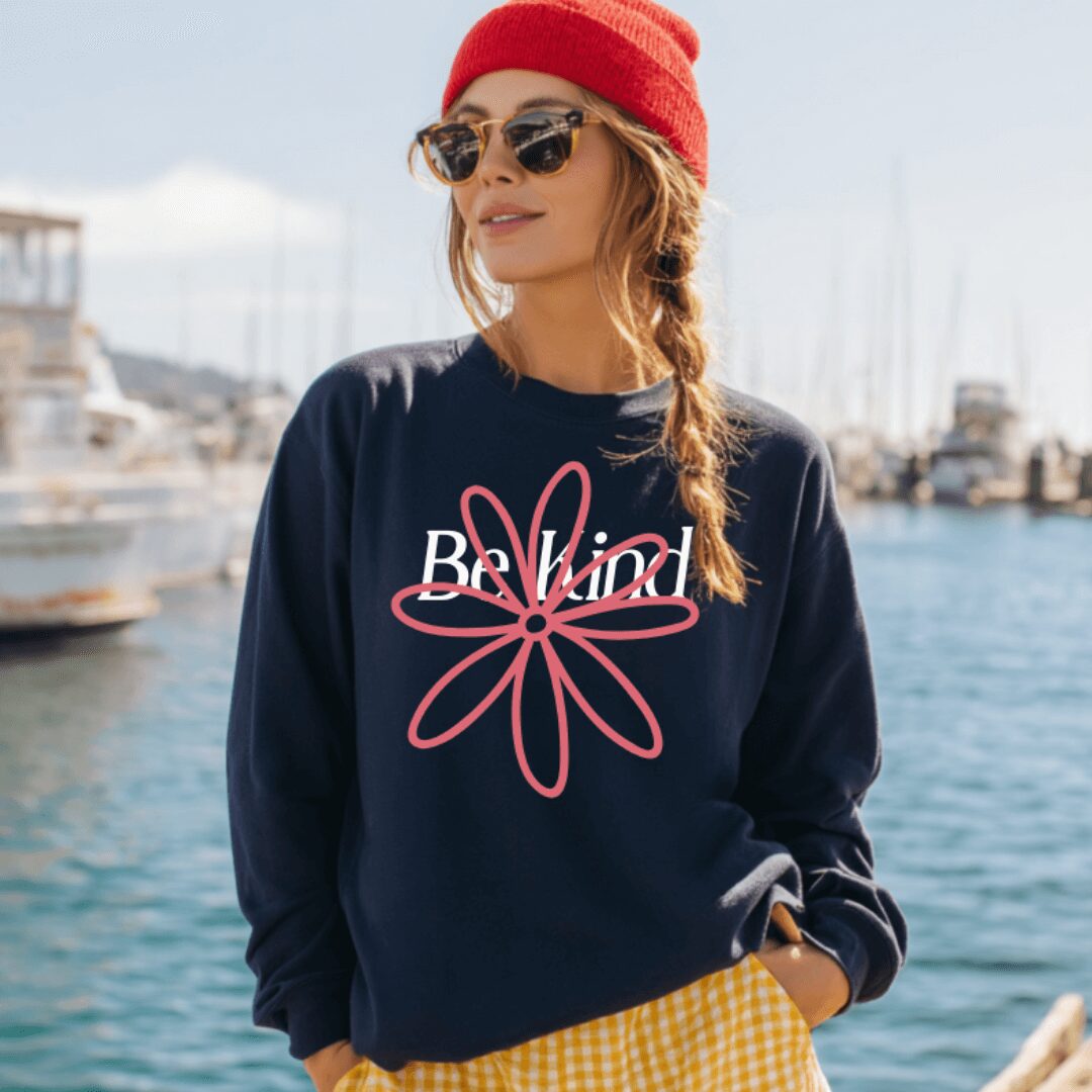 Woman wearing sunglasses, red beanie, navy sweatshirt with pink flower design, and yellow checkered pants, standing near boats and water.