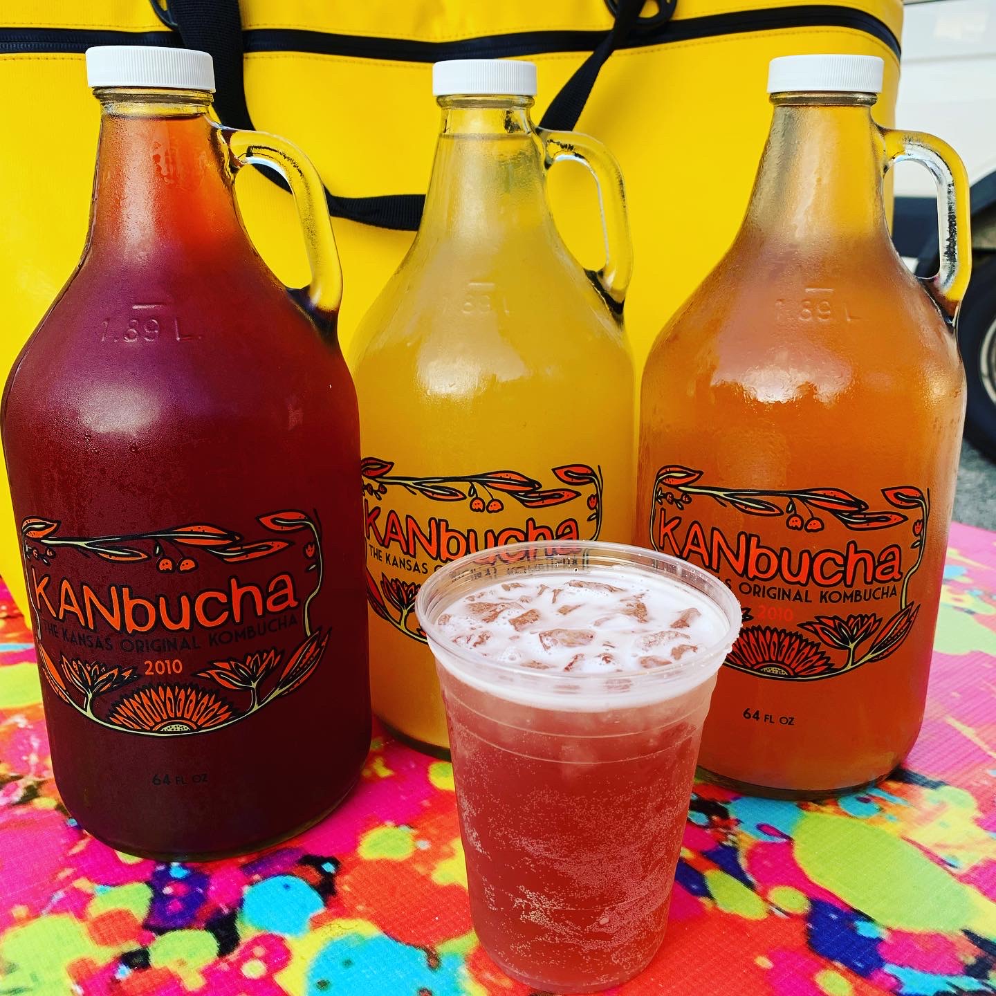 Three large bottles of kombucha and a glass of kombucha on a colorful tablecloth, with a yellow bag in the background.