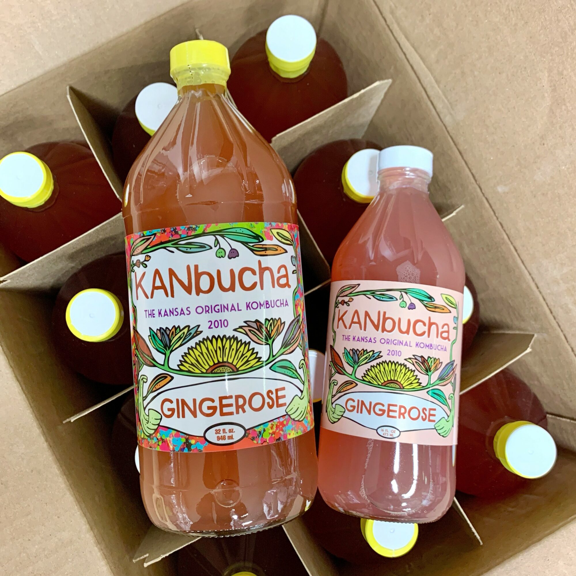 Two bottles of gingerose kombucha inside a cardboard box, with additional bottles in the background. The bottles have colorful labels.