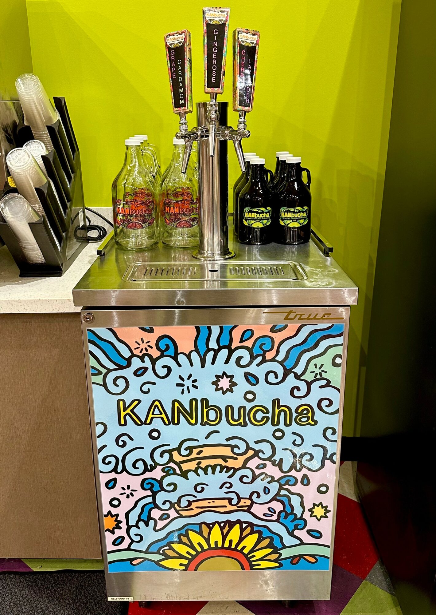 Kambucha dispenser with colorful, artistic design on front, set against a green wall, with bottles and utensils nearby.