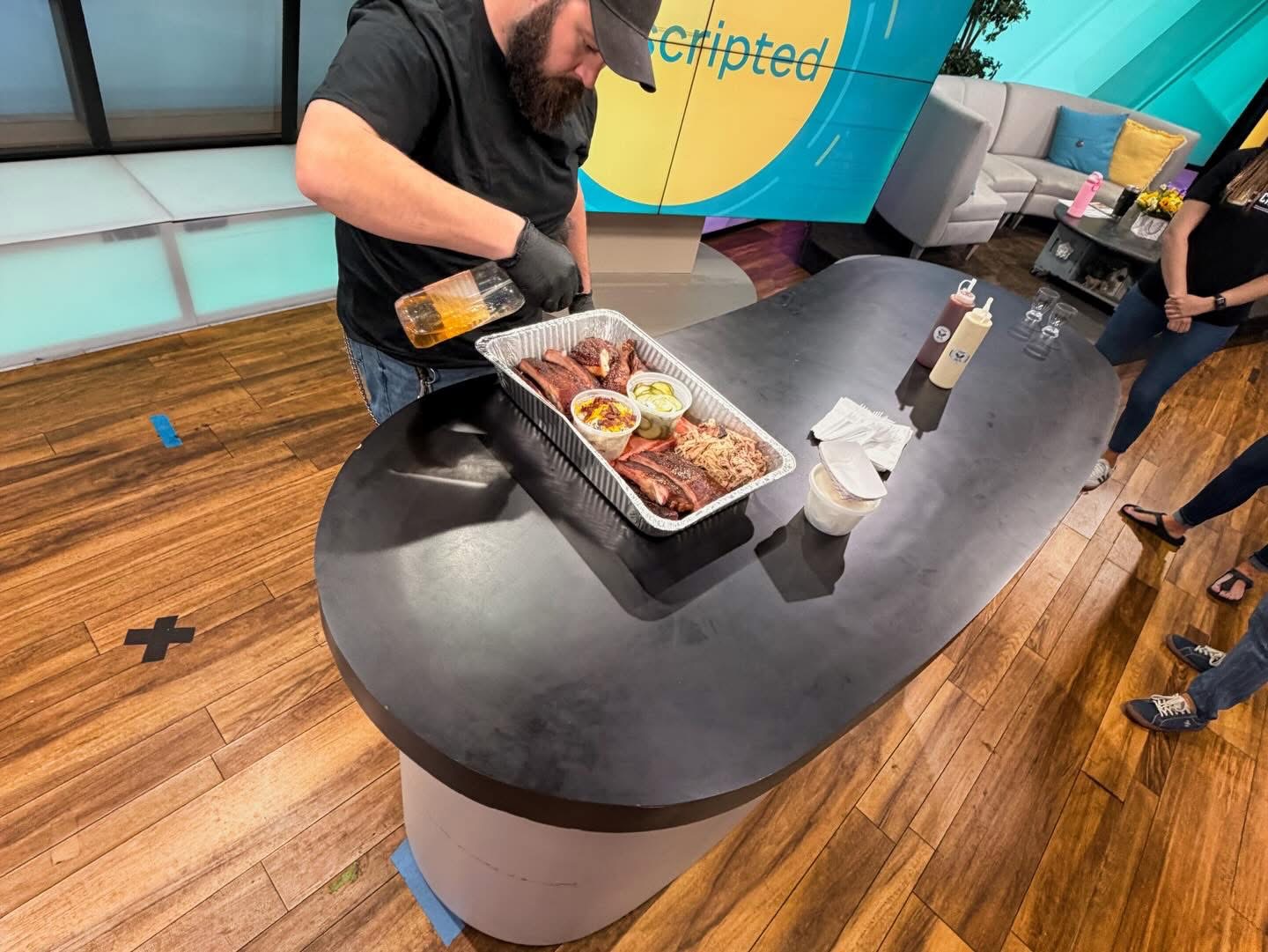Man preparing food at a curved black countertop with people in the background, wooden floor, and colorful wall.